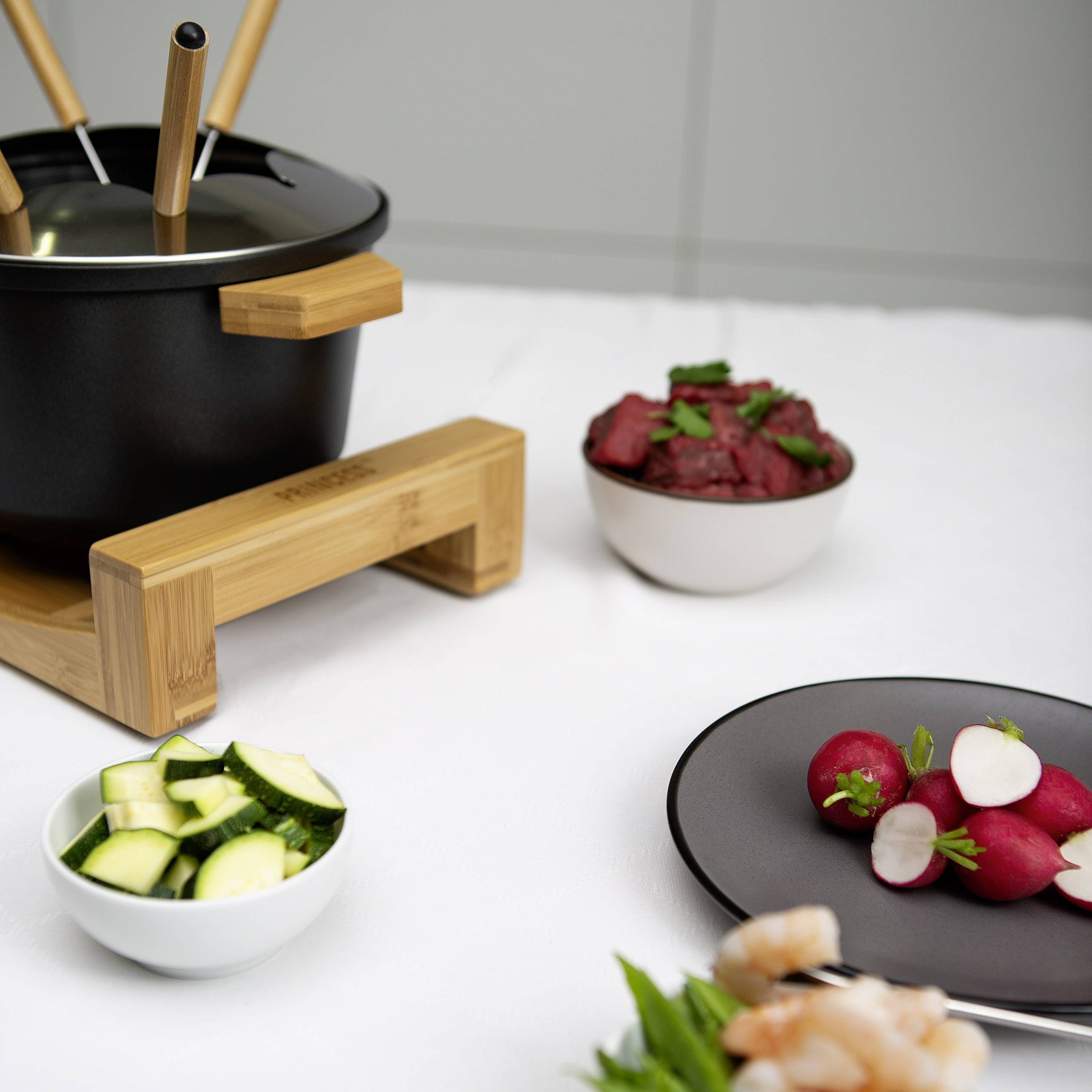 Un set à fondue avec support en bois est posé sur une table. À côté se trouvent des bols contenant des morceaux de courgettes, de betteraves et une assiette noire avec des radis.