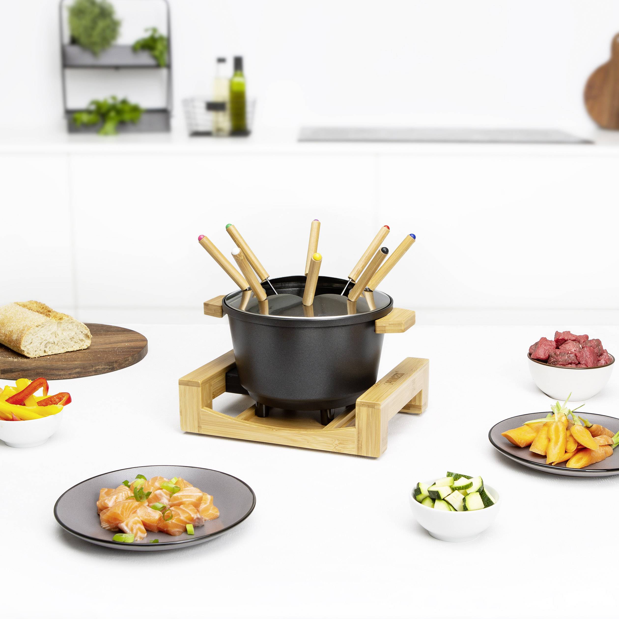 Un set à fondue avec un cadre en bois au centre, entouré d'assiettes de pain, de légumes et de viande, sur une table de cuisine blanche.
