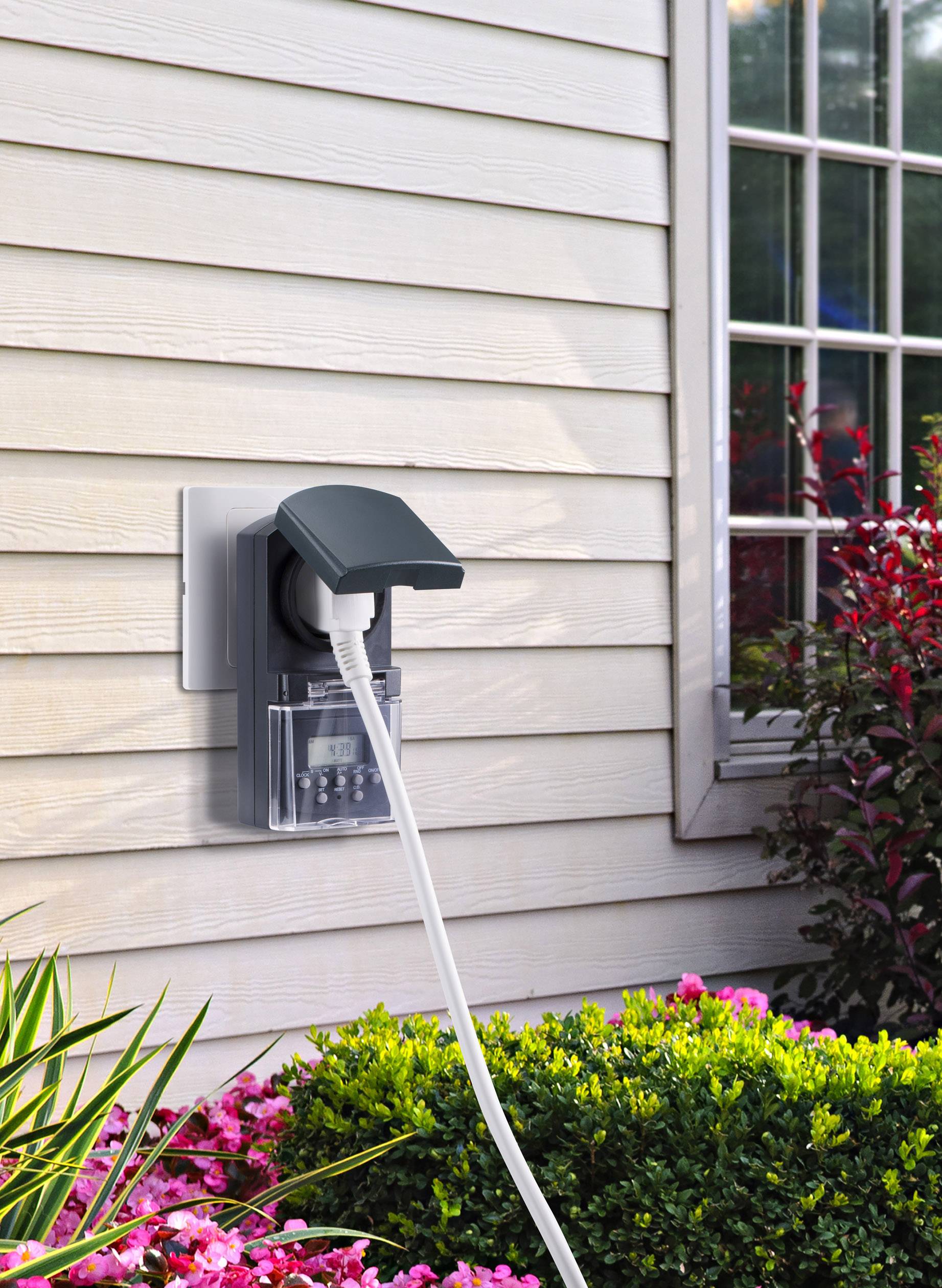 Prise électrique extérieure sur un mur de maison avec un couvercle de protection. Un câble est branché, à côté d'une fenêtre et de buissons.