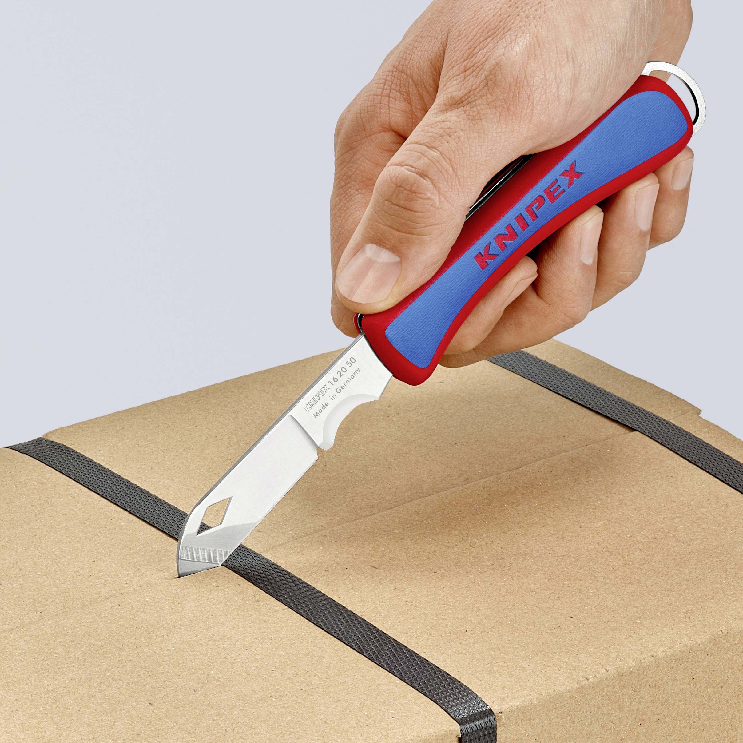 Une main tient un couteau à découper rouge et bleu portant l'inscription 'Knipex' et coupe un ruban noir sur un carton.