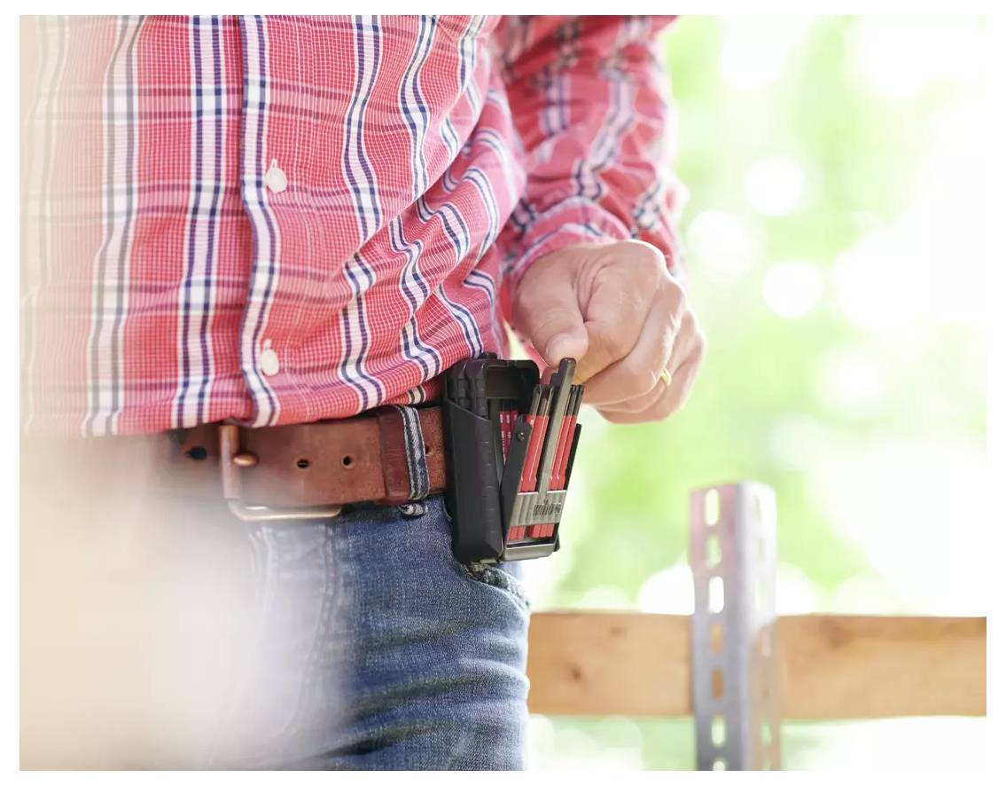 Une personne vêtue d'une chemise à carreaux rouge et d'un jean tient des crayons dans un étui de ceinture. L'arrière-plan présente une végétation floue, suggérant un cadre extérieur.