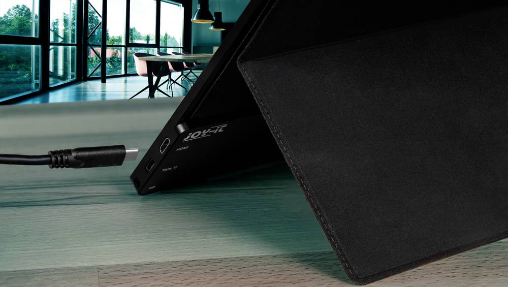 Un ordinateur portable est en train de se charger avec un câble de chargement sur une table en bois ; des grandes fenêtres et des meubles de bureau sont visibles en arrière-plan.