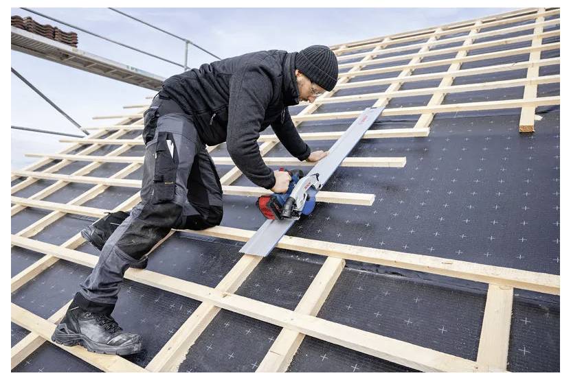 Un travailleur équipé de vêtements de protection de sécurité est à genoux sur la charpente en bois d'un toit, utilisant une scie circulaire pour couper une poutre horizontale. Des échafaudages sont visibles en arrière-plan.