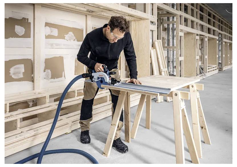 Une personne dans un atelier utilise un outil électrique pour couper une planche en bois sur un tréteau, entourée de charpente en bois non finie et de plaques de plâtre.