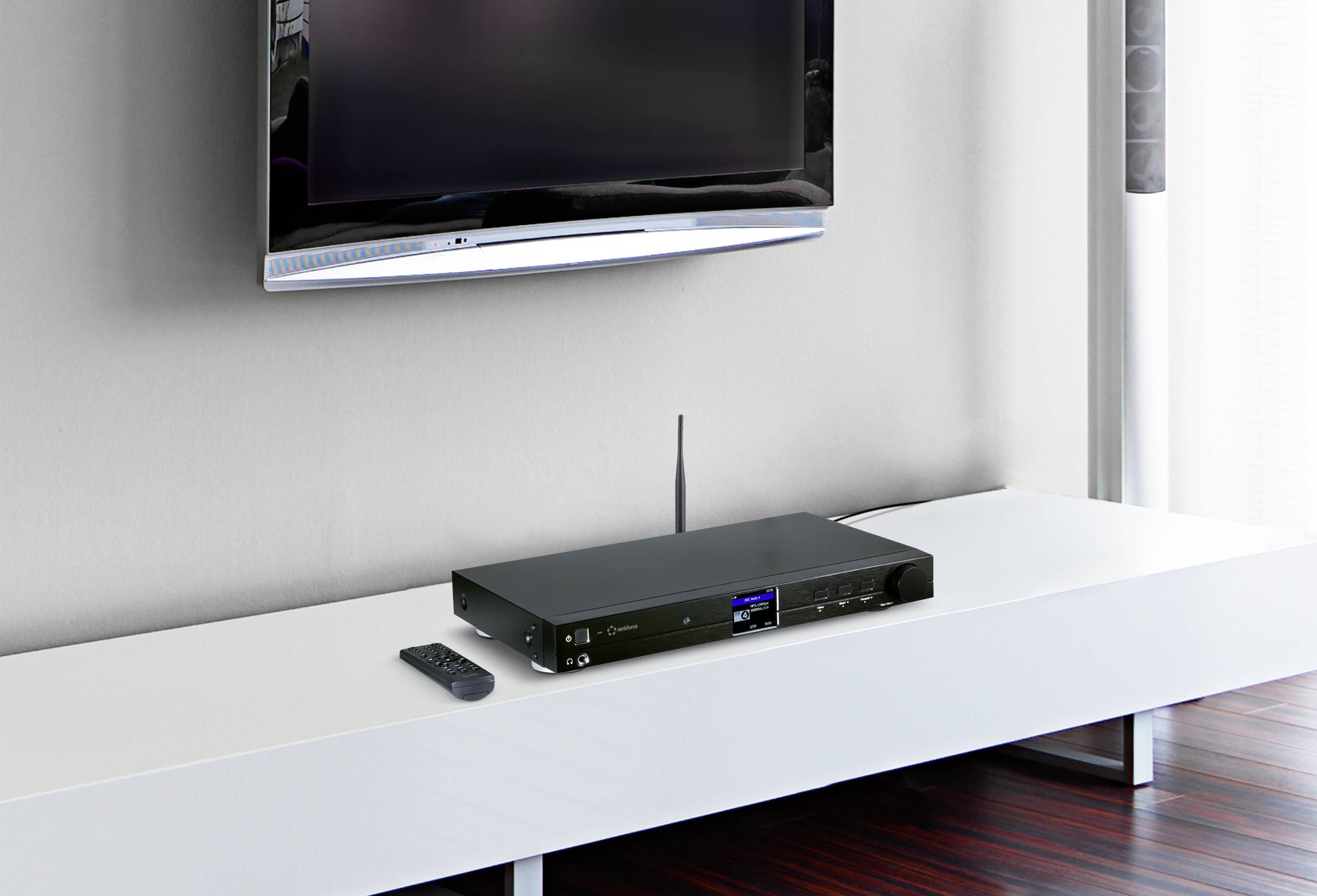Un récepteur noir moderne est posé sur une console TV blanche. Une télécommande est à côté. En arrière-plan, un téléviseur à écran plat est accroché au mur.