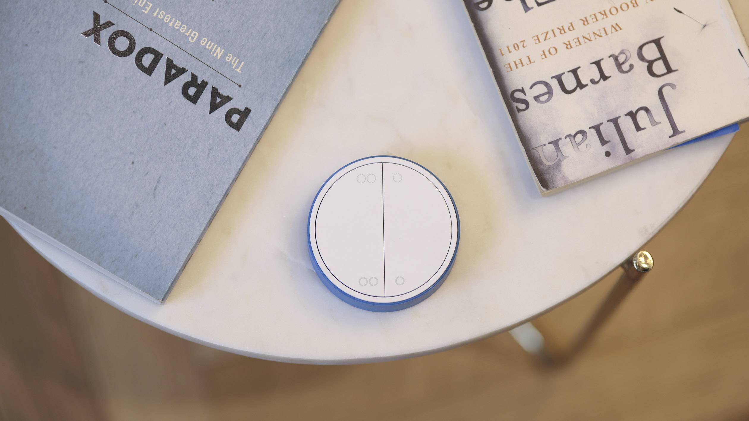 Un interrupteur rond blanc à deux boutons posé sur une table ronde à côté de deux livres aux couvertures bleues et beiges.