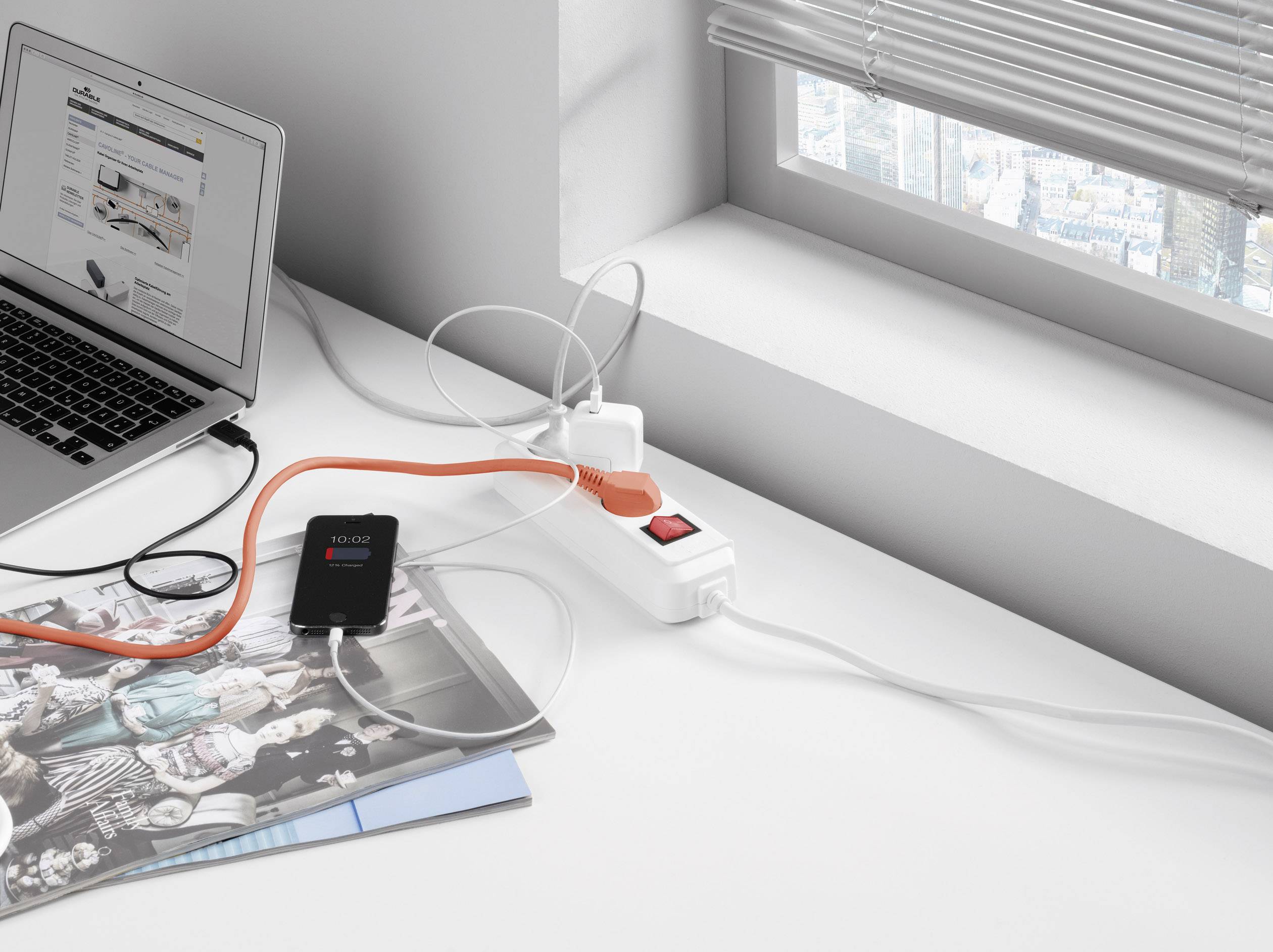 Un ordinateur portable avec une page web ouverte, branché à une multiprise avec plusieurs appareils, sur un bureau près de la fenêtre.