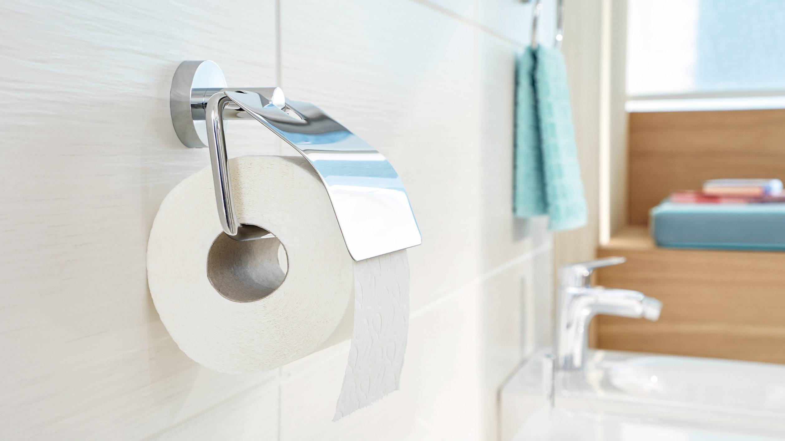 Un rouleau de papier toilette sur un support dans une salle de bain lumineuse. Un robinet et une serviette sont visibles en arrière-plan.