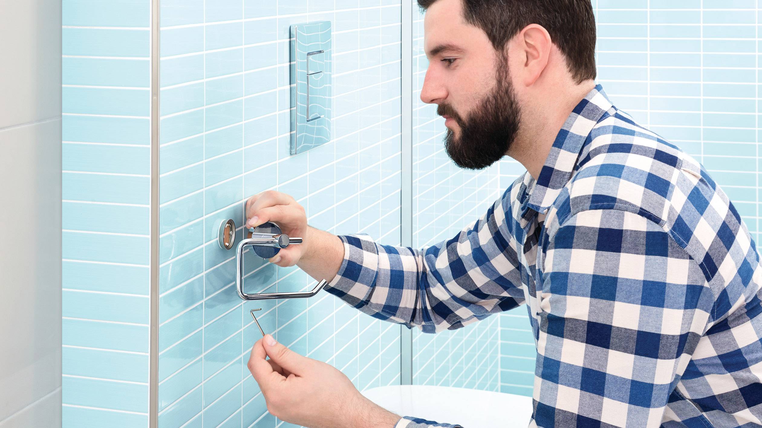 Un homme portant une chemise à carreaux installe ou répare un support de papier toilette sur un mur de salle de bain carrelé de bleu.