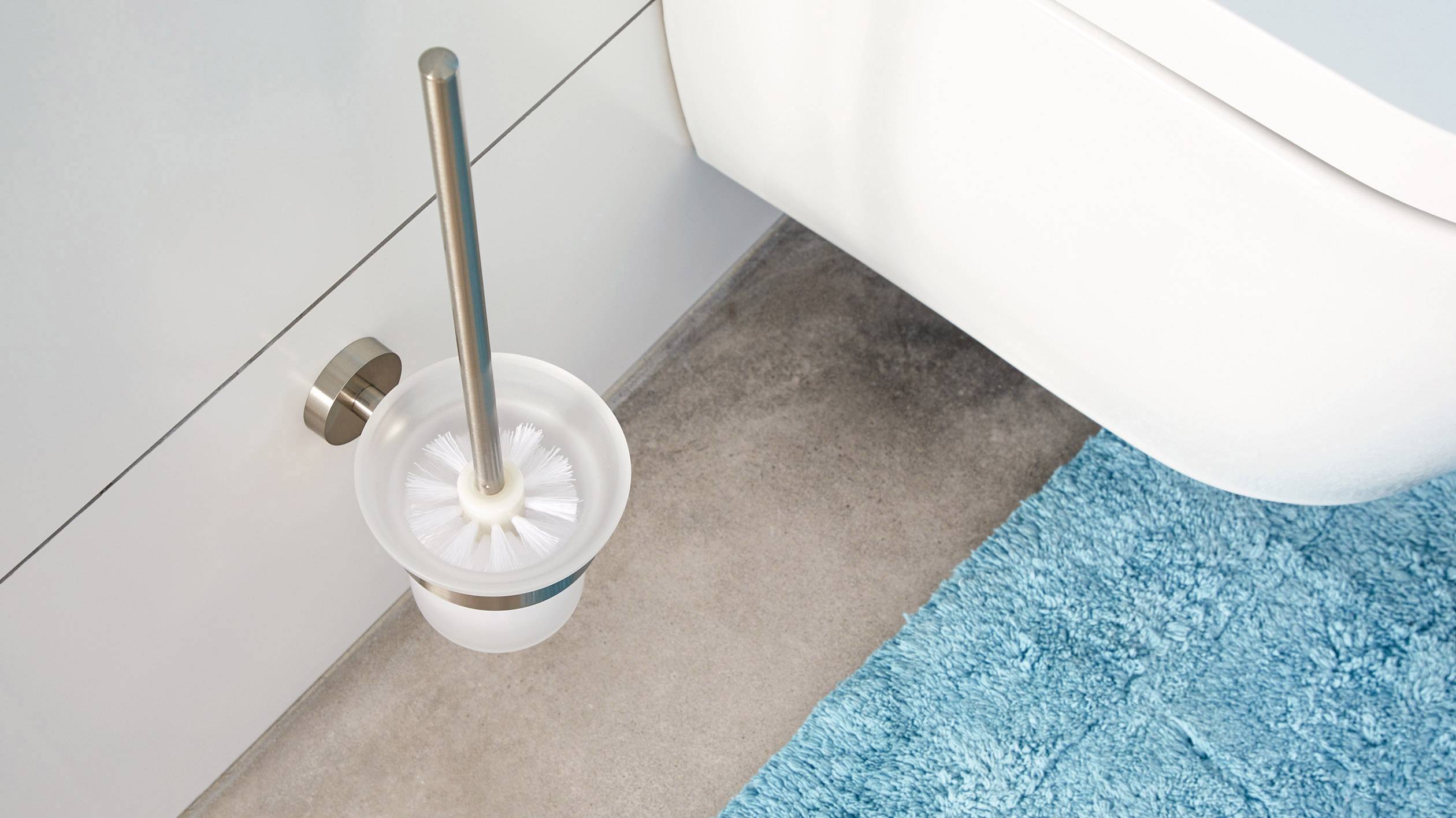Un support de brosse de toilette avec une brosse à l'intérieur est placé à côté des toilettes blanches sur un sol gris, avec un tapis bleu à proximité.
