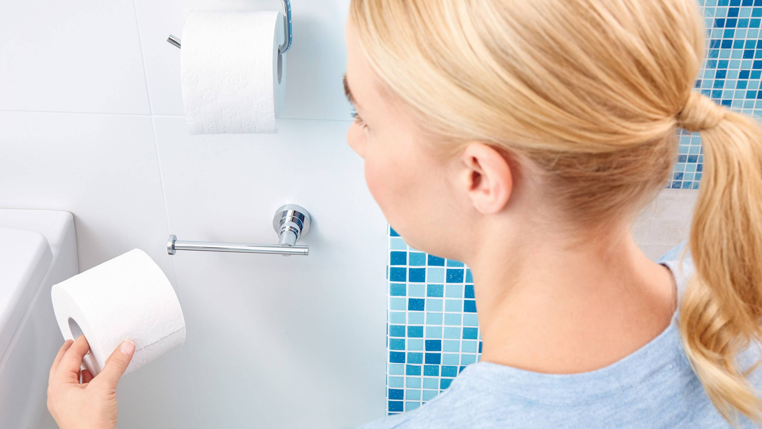 Une personne dans la salle de bain tient un rouleau de papier toilette tout en regardant un support mural avec un autre rouleau.