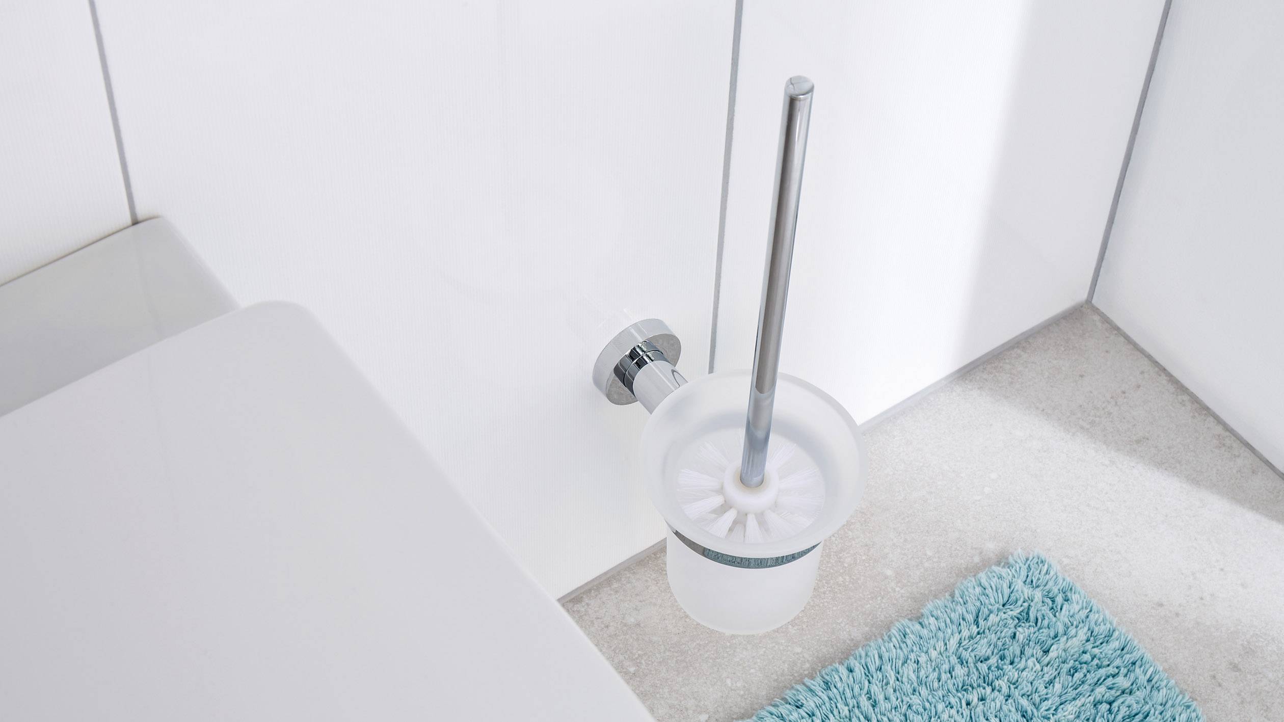 Brosse de toilette dans son support en plastique, posée sur le sol de la salle de bain à côté des toilettes et d'un tapis de bain bleu.