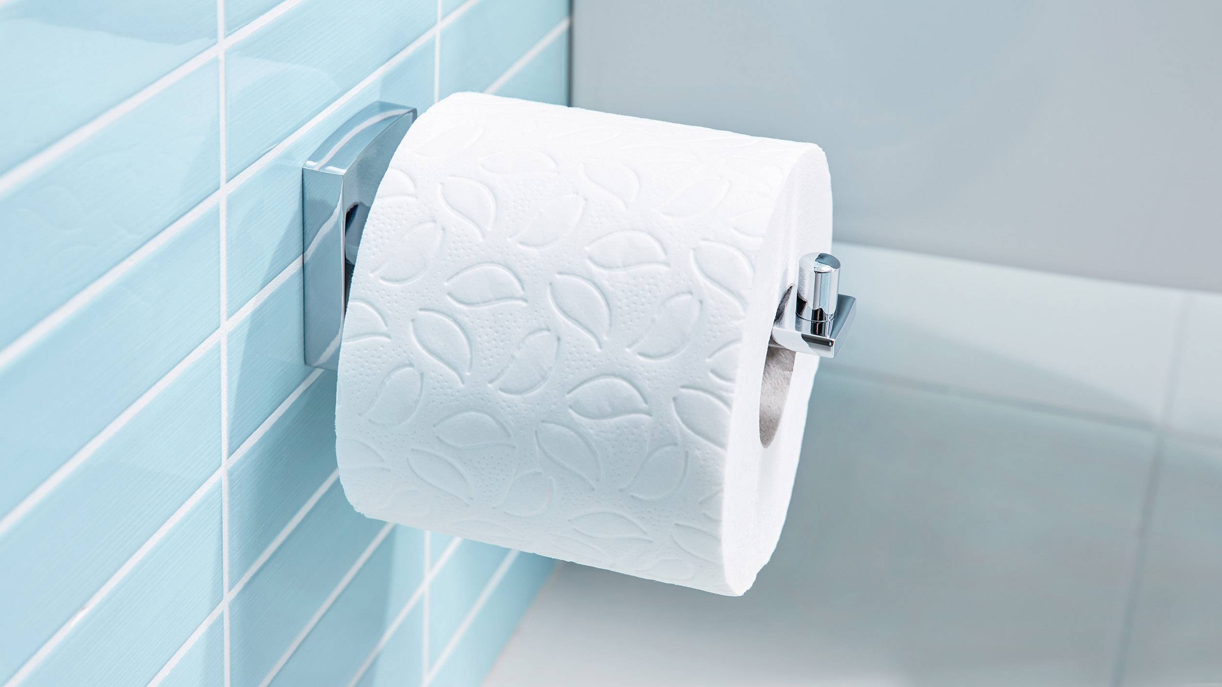 Rouleau de papier toilette sur un support en acier inoxydable, fixé sur un mur carrelé de couleur turquoise.