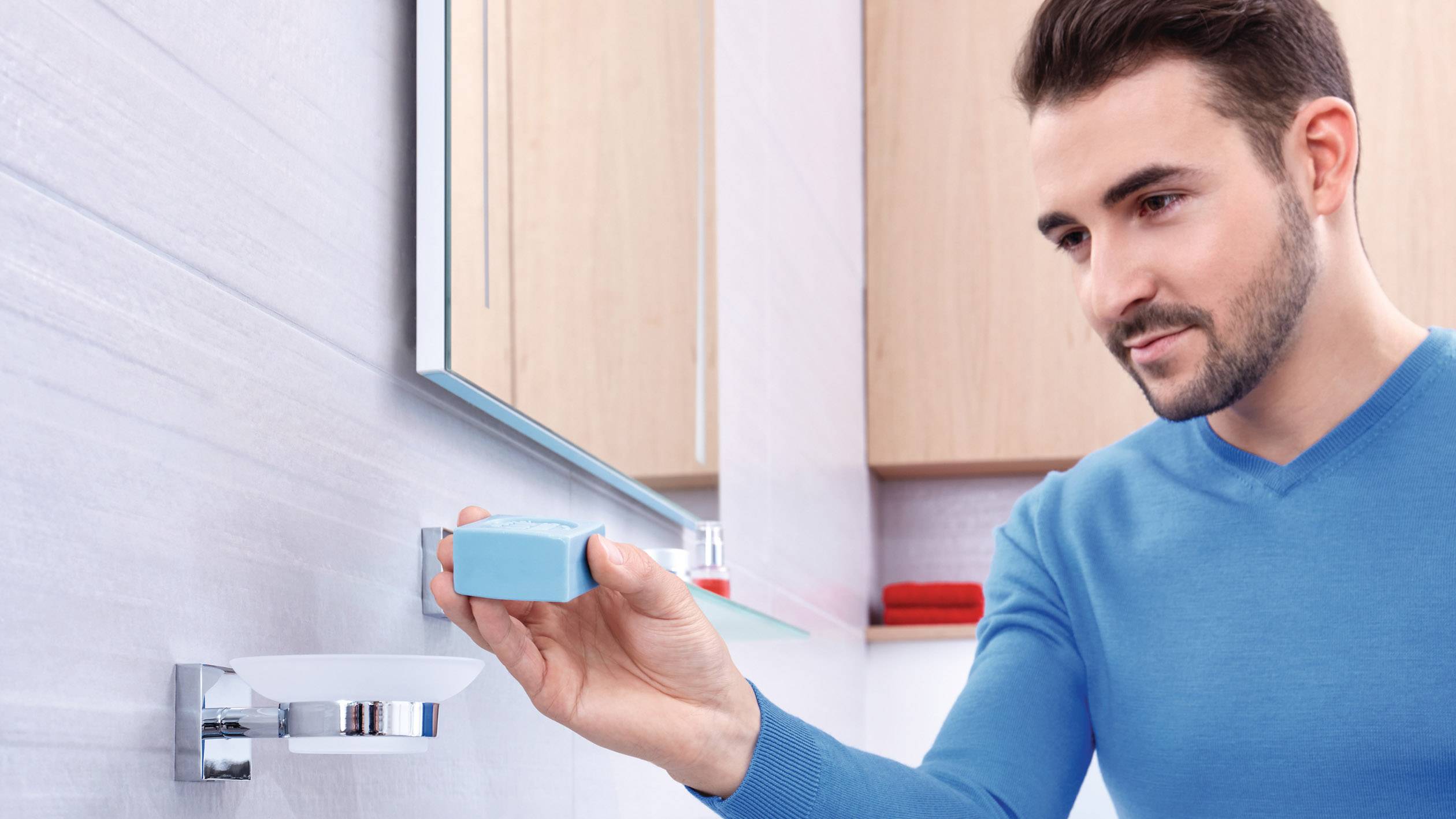 Un homme portant un pull bleu pose un morceau de savon bleu sur un porte-savon dans la salle de bain.