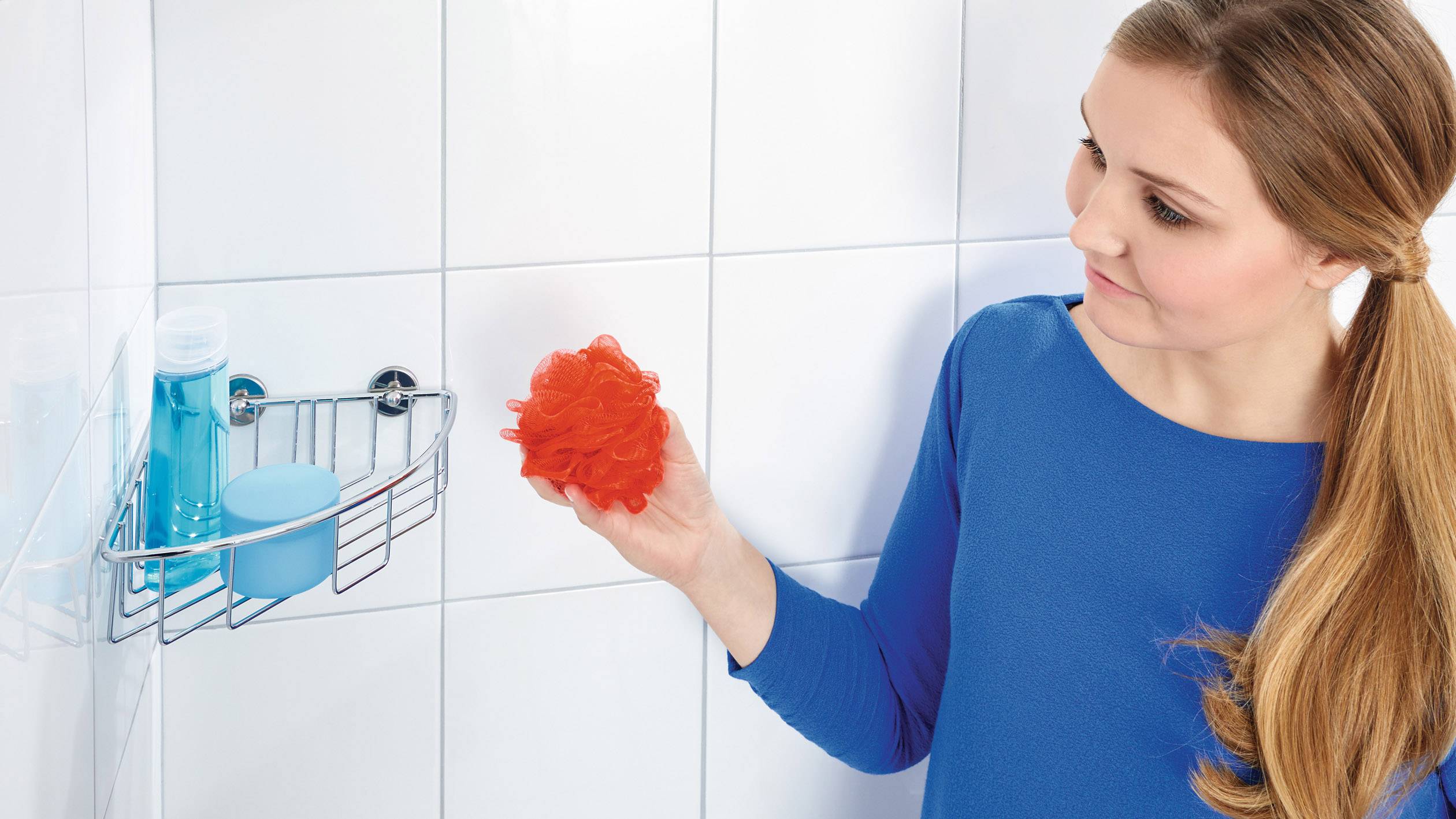 Une femme vêtue de bleu tient une éponge de douche orange à la main, debout devant une étagère de douche avec une bouteille de savon bleue et un récipient.