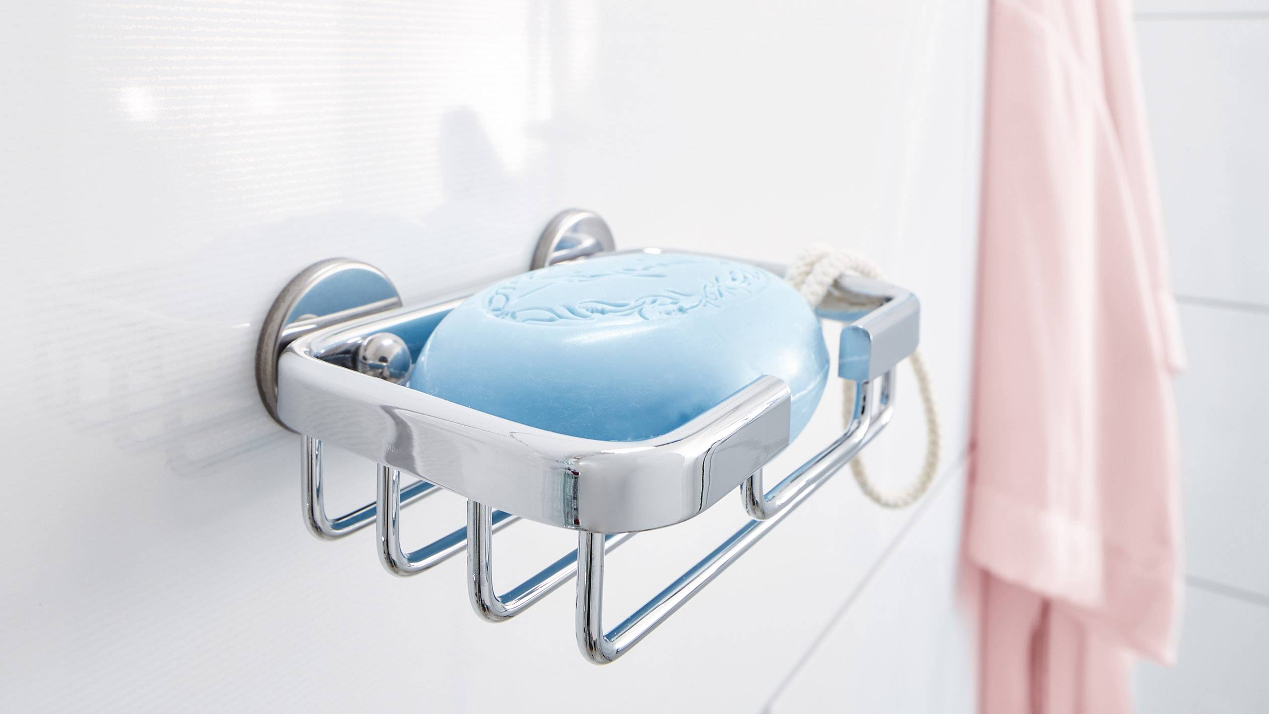 Morceau de savon bleu dans un support métallique sur un mur blanc, avec un peignoir rose en arrière-plan. Accessoires de salle de bain.