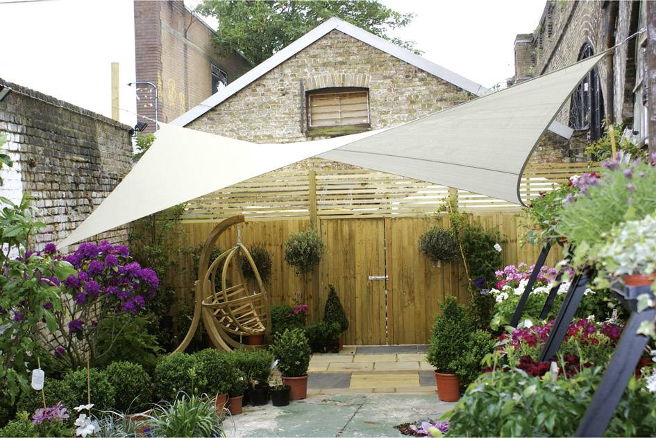 Un petit jardin de cour arrière avec des fleurs, des plantes et une chaise à bascule en bois sous un auvent en tissu ressemblant à une voile de soleil.