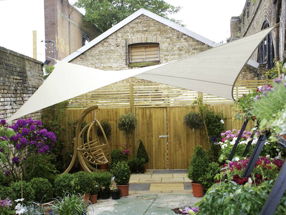 Un petit jardin avec de nombreuses plantes en pot, un balançoire en bois et des voiles solaires. Maison de briques en arrière-plan.