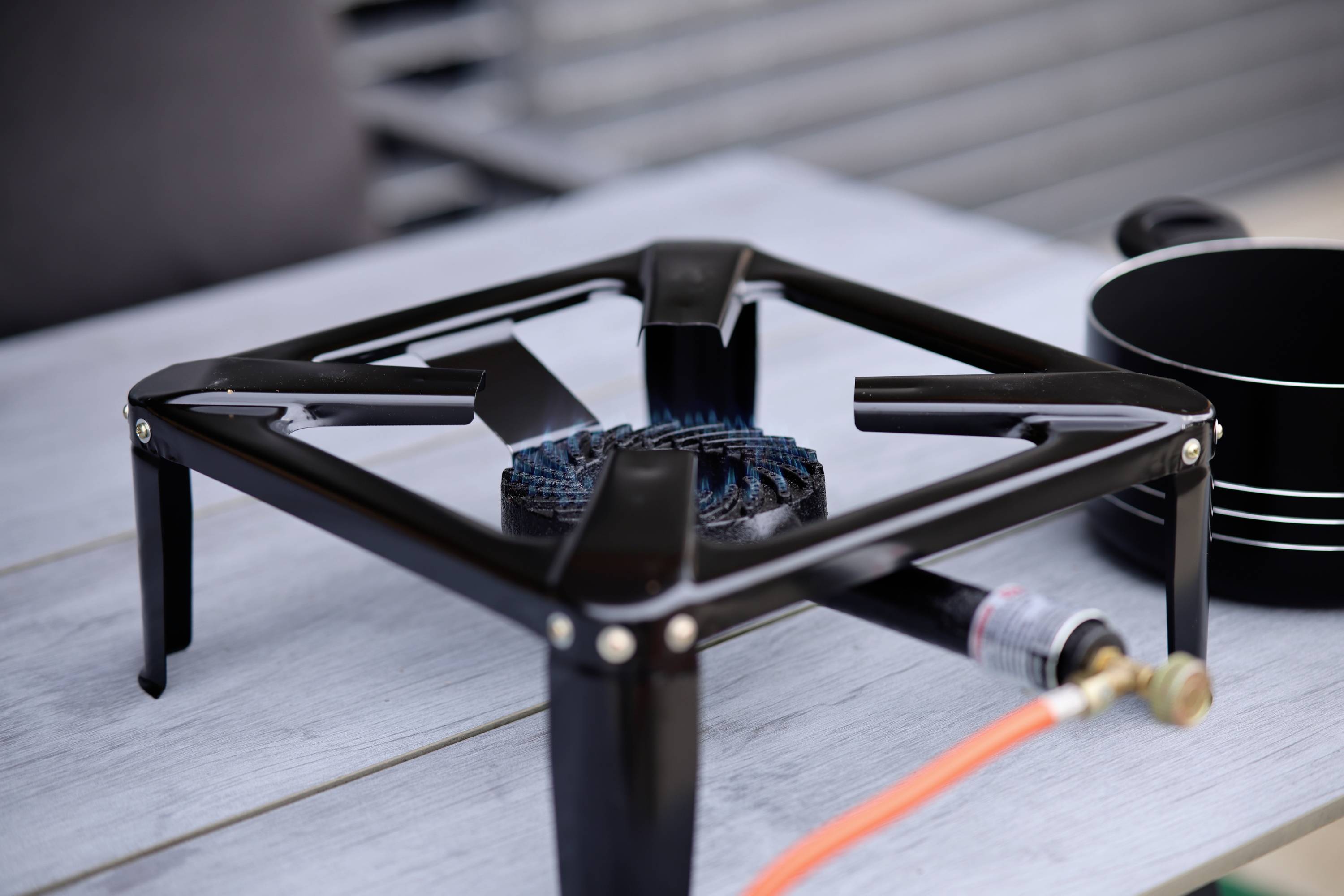 Un brûleur à gaz noir avec un tuyau orange est posé sur une table grise. À côté se trouvent des casseroles noires.