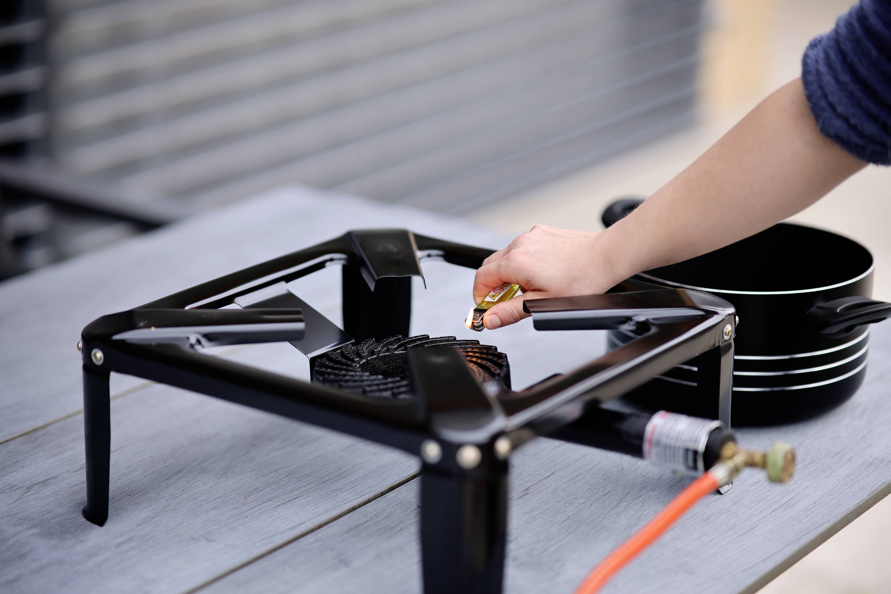 Une main tourne un tuyau de gaz sur un réchaud de camping noir posé sur une table en bois à l'extérieur. Une casserole est prête à être utilisée.