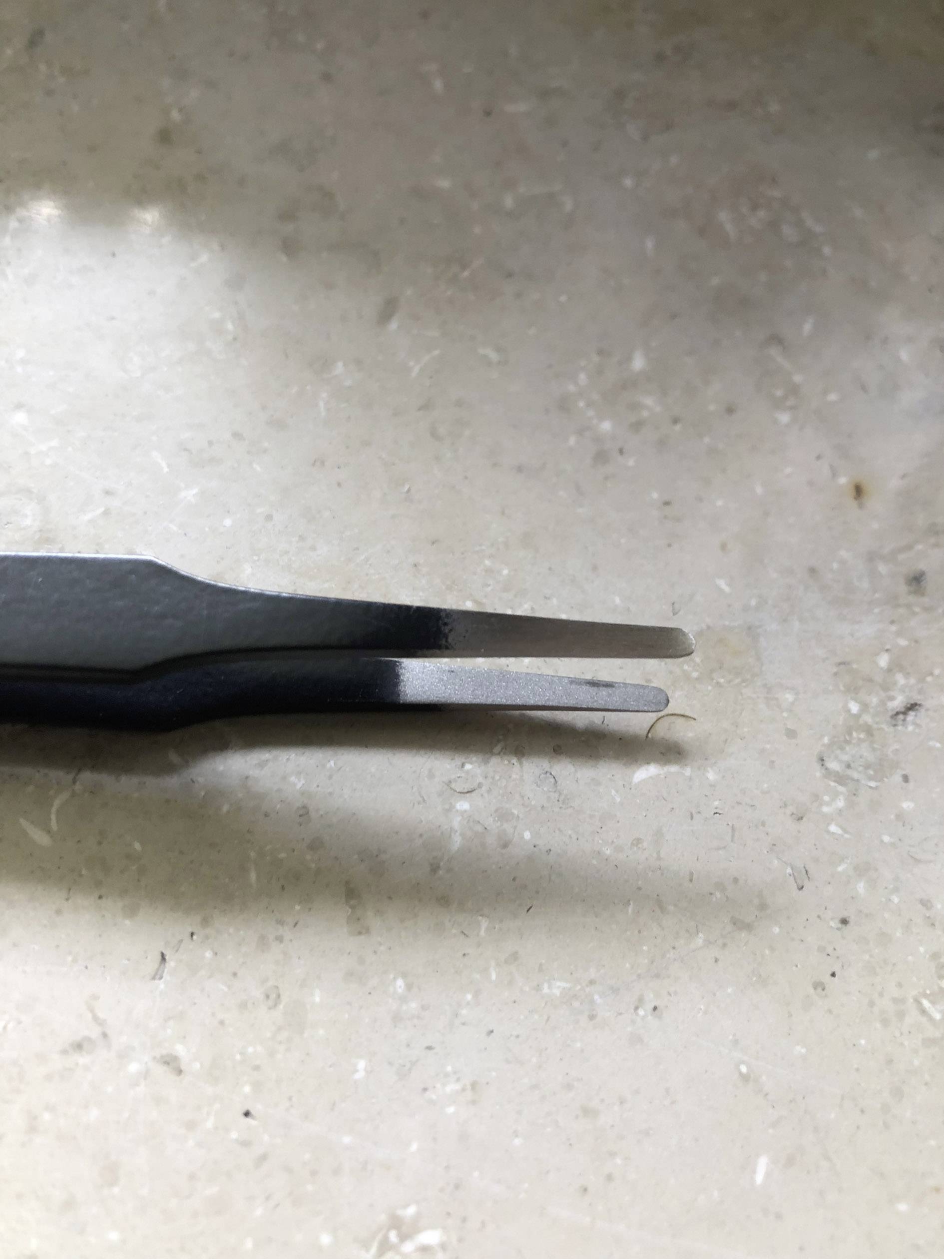 Une pince à épiler repose sur une surface de pierre claire. Ses pointes sont légèrement incurvées et ne se rejoignent pas tout à fait.