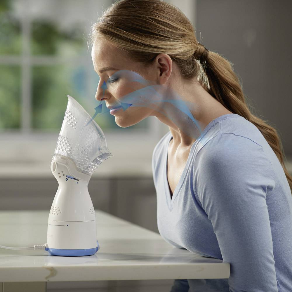 Une femme utilise un inhalateur électrique pour traiter les voies respiratoires. La flèche bleue indique le trajet de l'air depuis l'appareil jusqu'au nez.