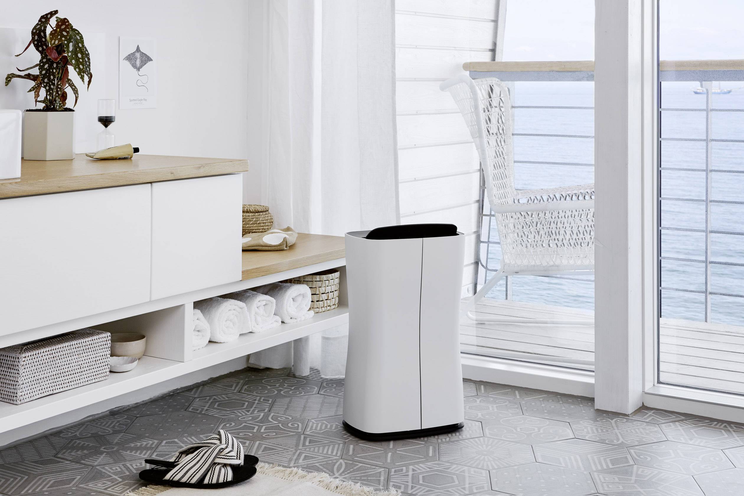 Purificateur d'air blanc sur un sol gris dans une pièce moderne, avec vue sur la mer à travers de grandes fenêtres, entouré de meubles blancs.