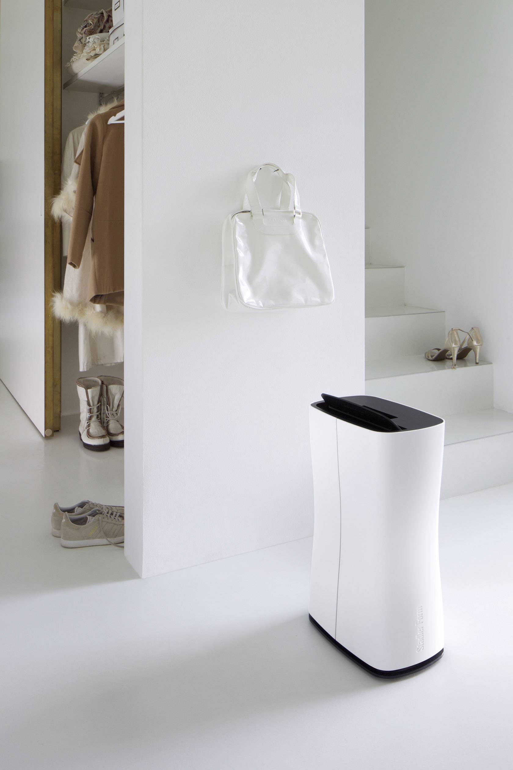 Un purificateur d'air blanc est placé devant un mur blanc dans une pièce moderne et minimaliste, avec des chaussures et un sac à main.