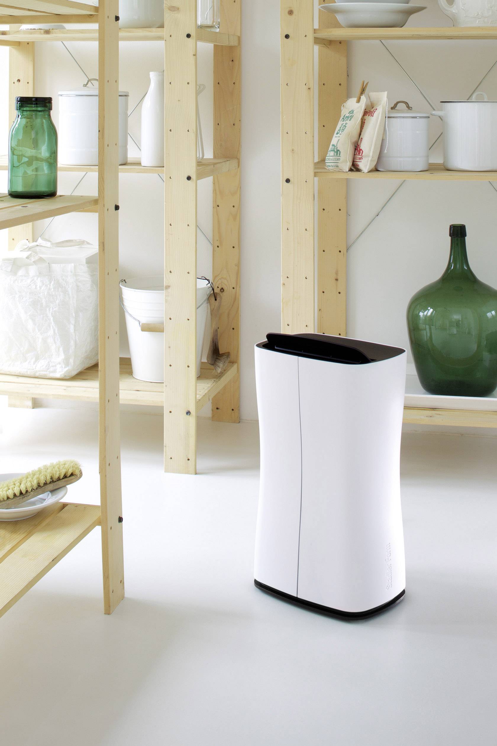 Un purificateur d'air blanc est posé sur un sol blanc, entre des étagères en bois dans une cuisine. Au fond, on aperçoit des bocaux et des contenants de stockage.