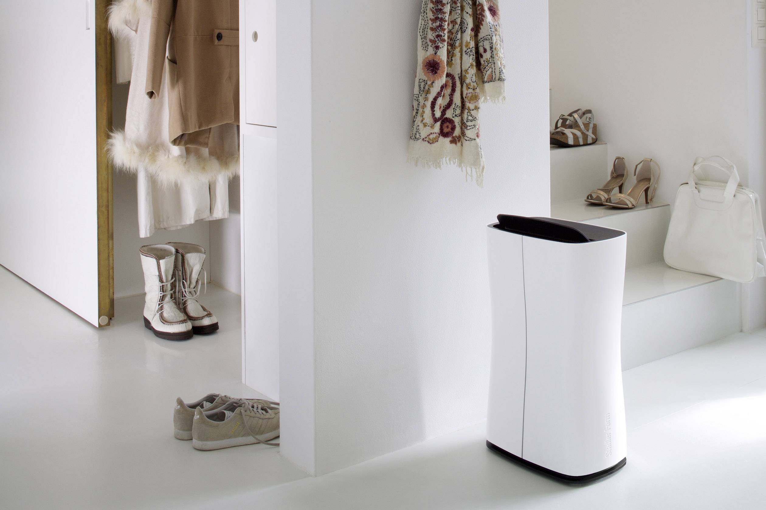 Un purificateur d'air élégant et blanc est placé dans un hall d'entrée moderne et sophistiqué. À côté se trouvent des bottes, des chaussures et un manteau.