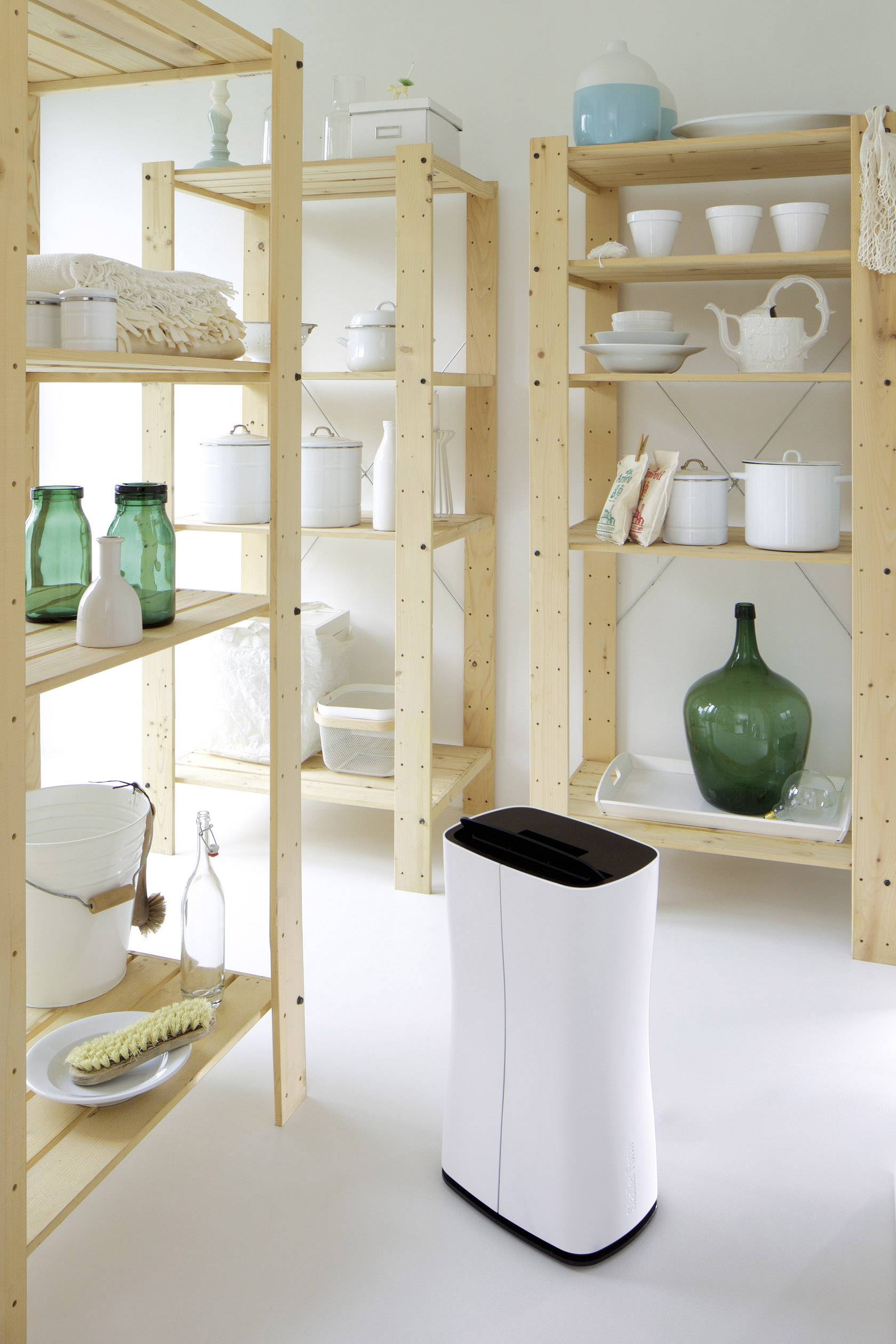 Un déshumidificateur blanc est placé dans une pièce avec une étagère en bois, contenant de la céramique, des bouteilles en verre et des objets ménagers.