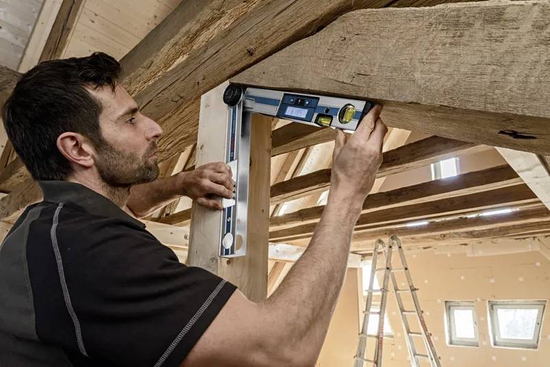 Une personne utilise un rapporteur d'angle numérique sur une poutre en bois dans une pièce en cours de construction, illustrant la précision du travail de charpenterie.