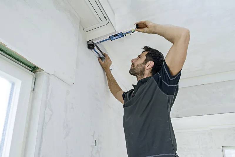 Un homme installe une unité de climatisation sur un mur à l'intérieur d'une pièce. Il utilise un outil pour fixer l'unité en place.
