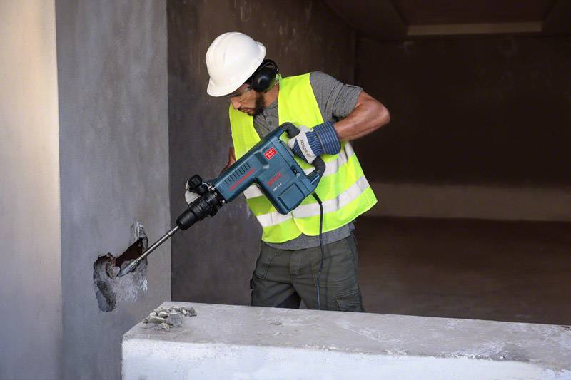 Un ouvrier portant un casque de chantier et un gilet de sécurité fore un trou dans un mur en béton à l'aide d'un outil électrique lourd dans une pièce en cours de construction.