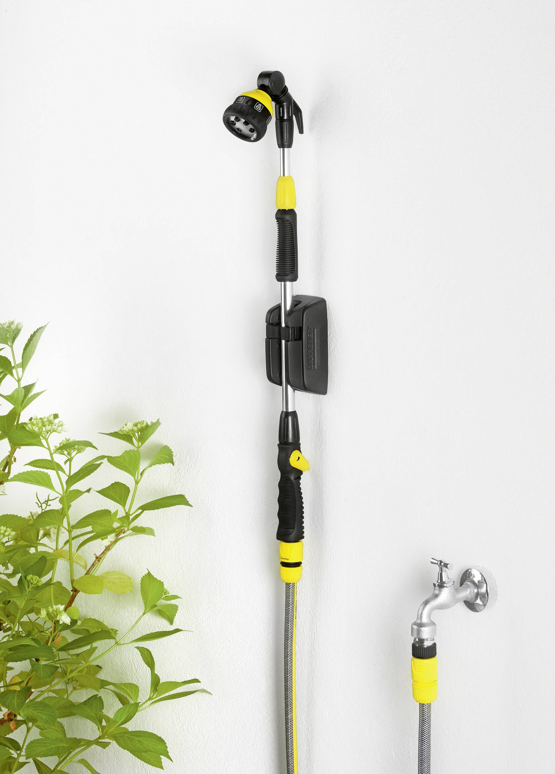 Pommeau de douche avec une poignée jaune et noire fixé à un mur. Un robinet est placé à droite et une branche de plante verte se trouve à gauche.
