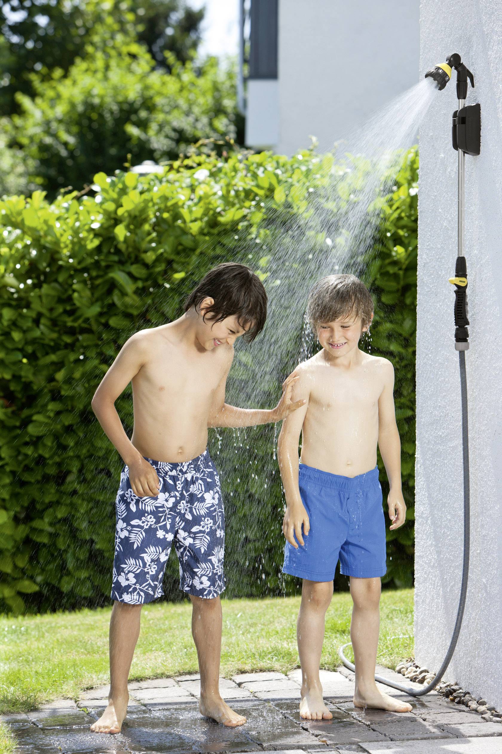 Deux garçons jouent à côté d'une douche extérieure dans un jardin. L'un asperge l'autre d'eau. C'est une journée ensoleillée.