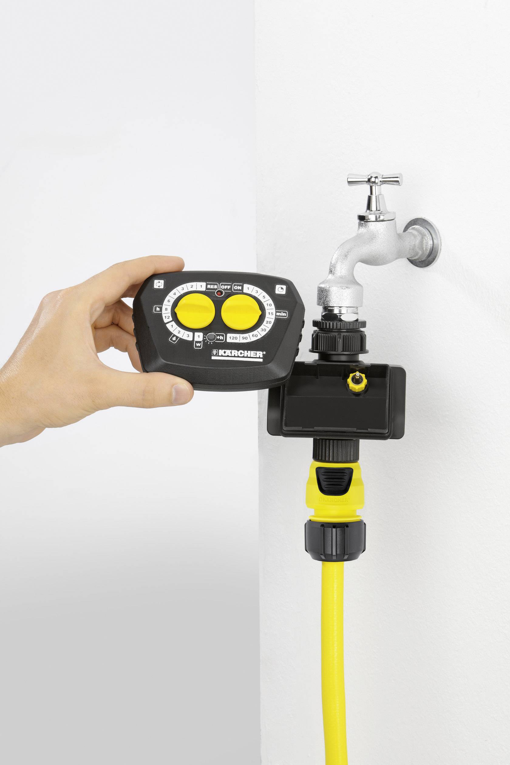 Une main tient un dispositif de commande d'irrigation devant un robinet avec un raccord de tuyau jaune, prêt à être installé.