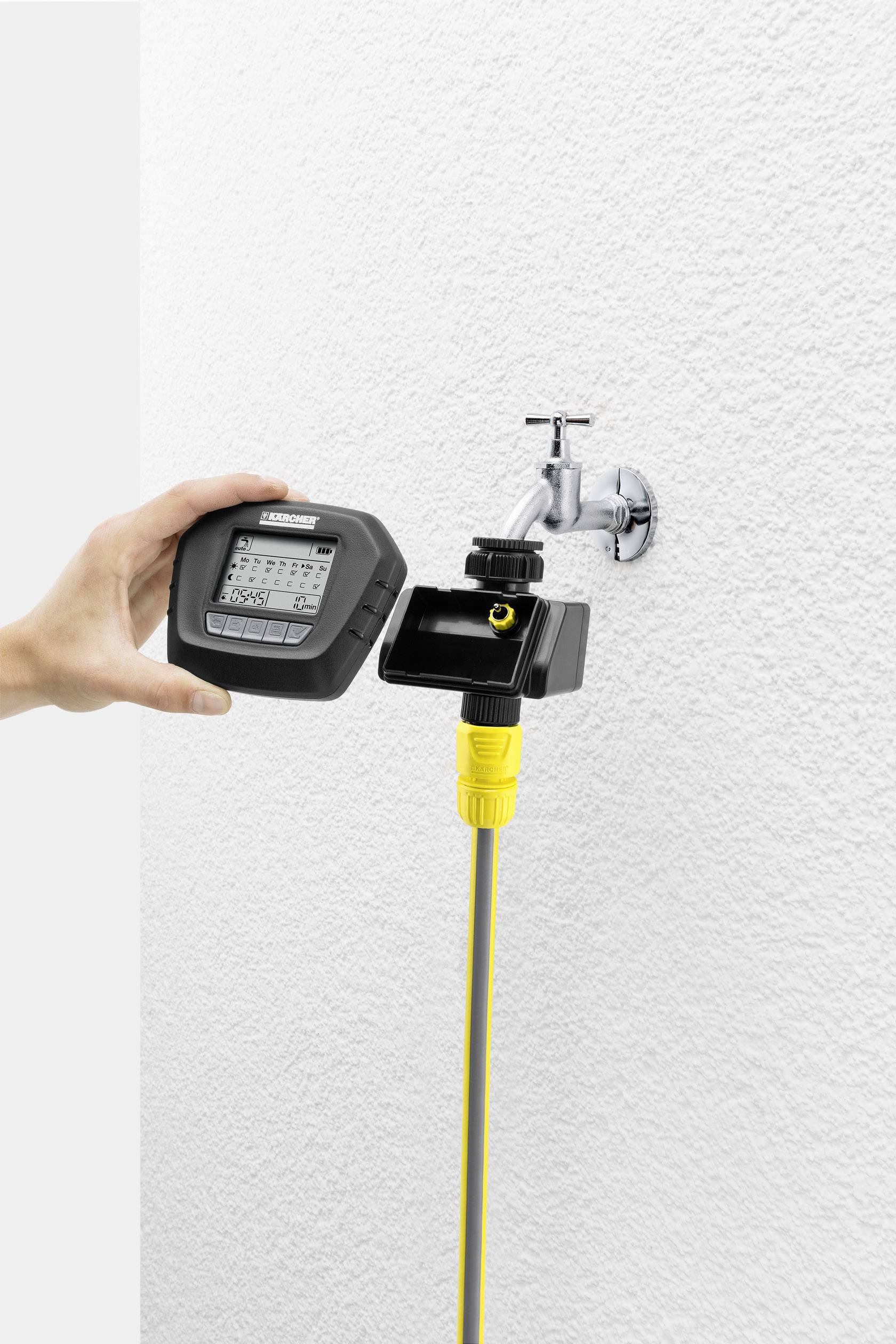 Une main tient un dispositif de contrôle d'irrigation à côté d'un robinet avec un tuyau jaune raccordé, contre un mur blanc.