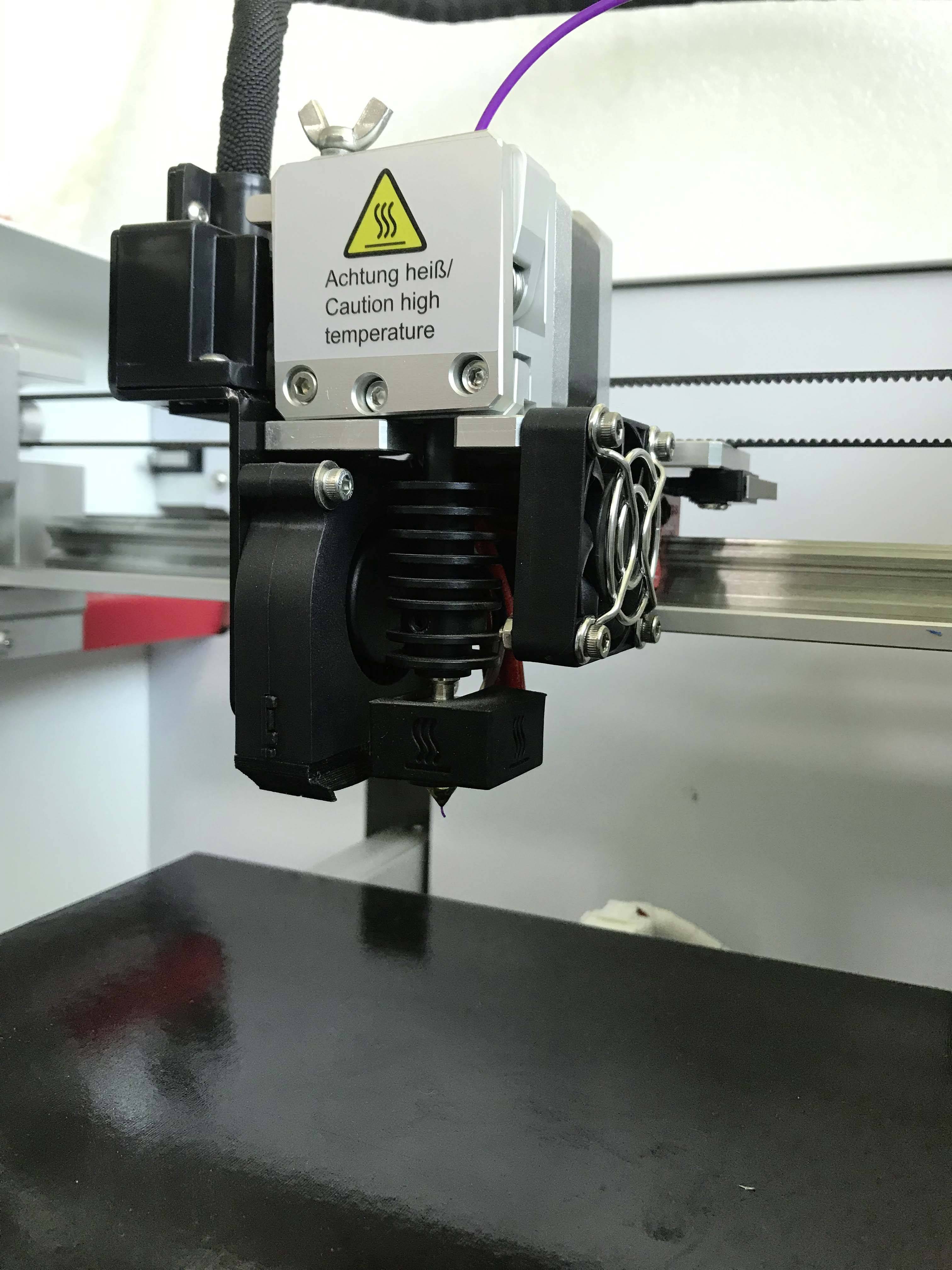 Imprimante 3D avec panneau d'avertissement 'Attention chaud !/Caution high temperature'. Gros plan de la tête d'impression en cours de fonctionnement.