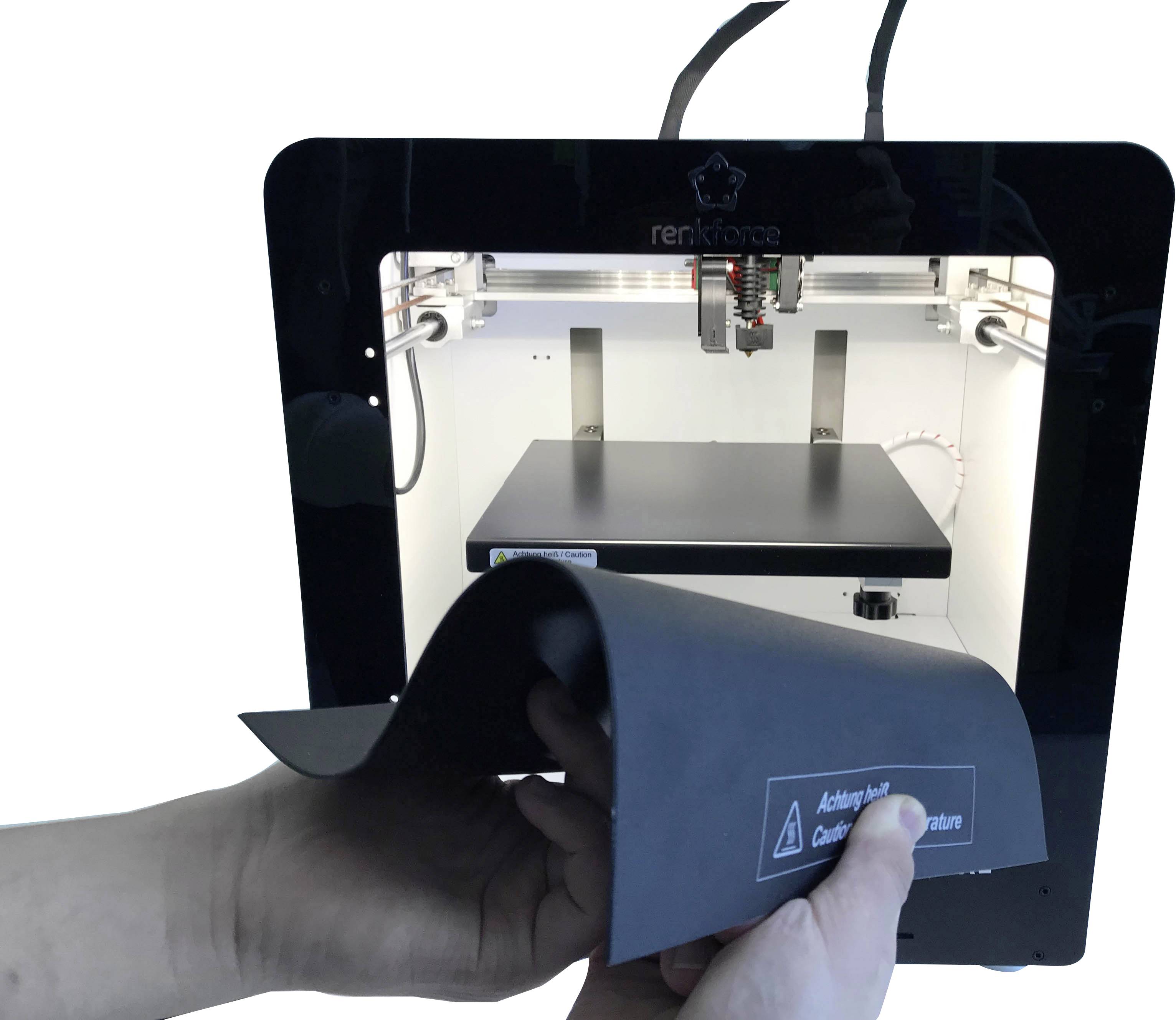 Une imprimante 3D imprime un objet en plastique. Des mains tiennent une plaque d'impression flexible avec un avertissement de sécurité.
