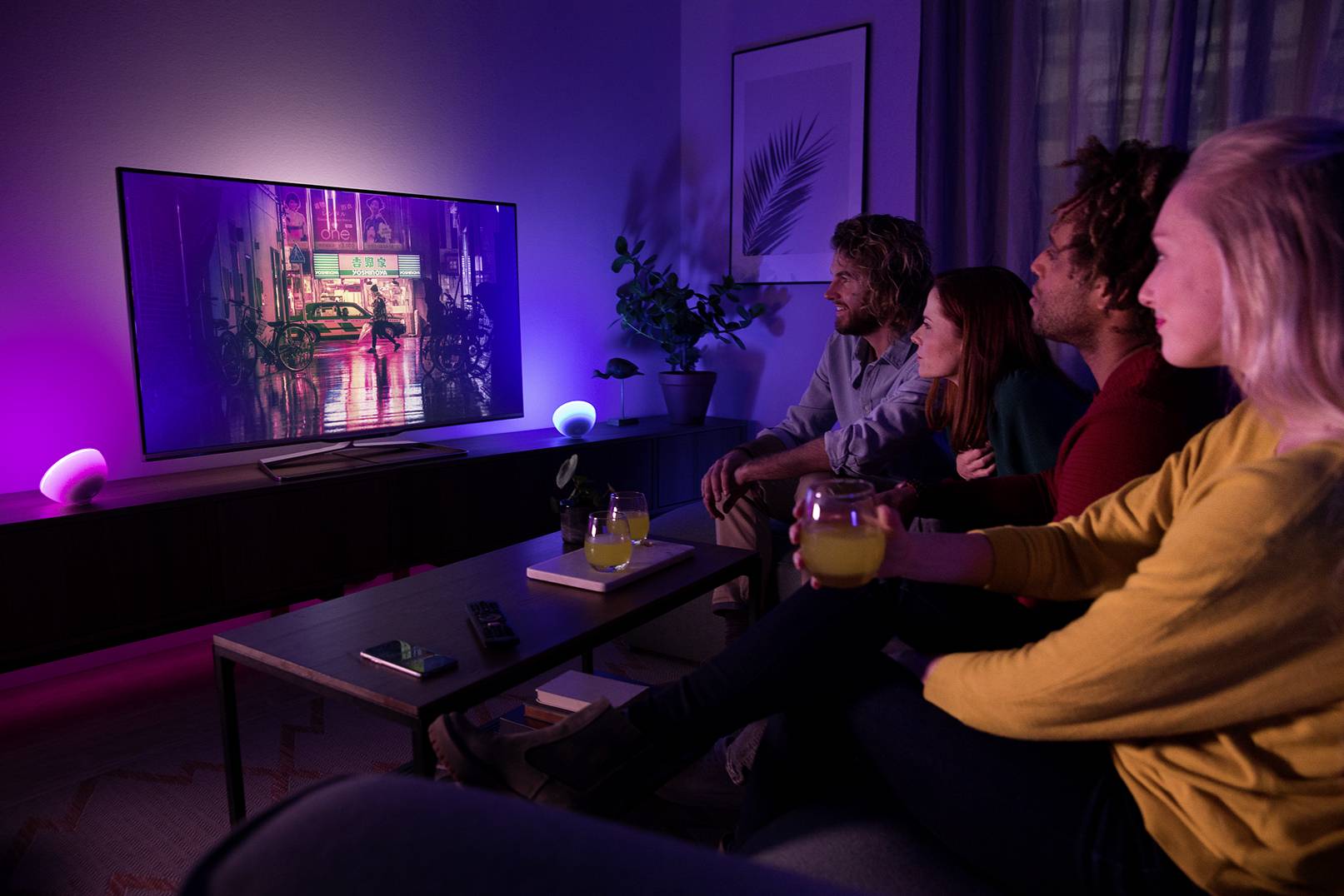 Un groupe de quatre personnes est assis dans un salon tamisé, regardant un grand téléviseur avec un éclairage de fond coloré.
