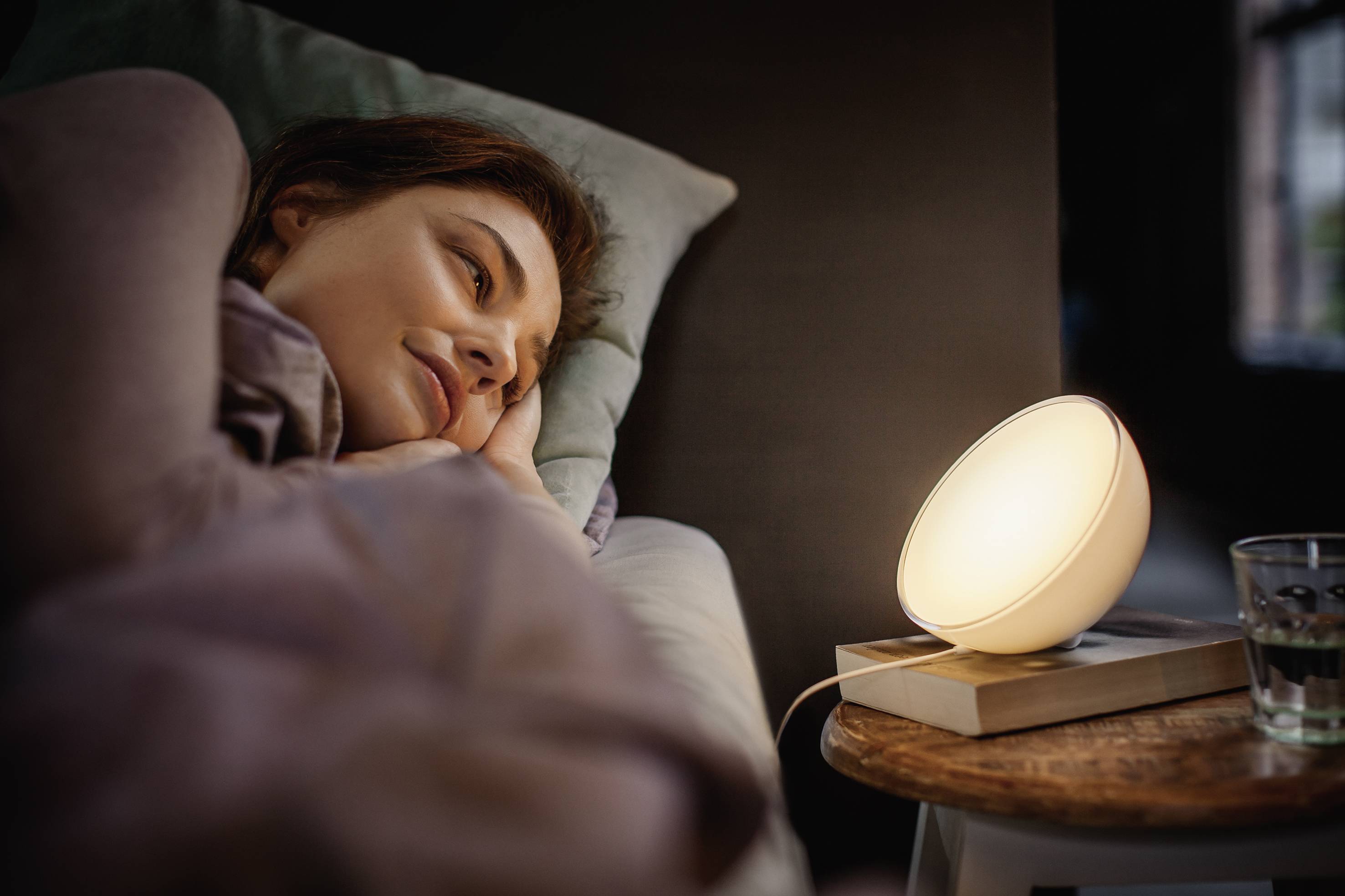 Une femme est allongée dans son lit, regardant une lampe lumineuse sur la table de chevet. L'éclairage atmosphérique suggère la détente.