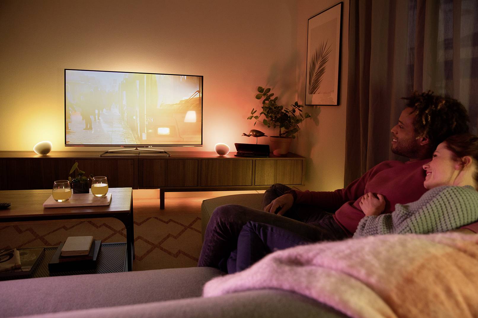 Un couple est confortablement installé sur le canapé et regarde la télévision. La pièce est doucement éclairée. Deux boissons sont posées sur la table.