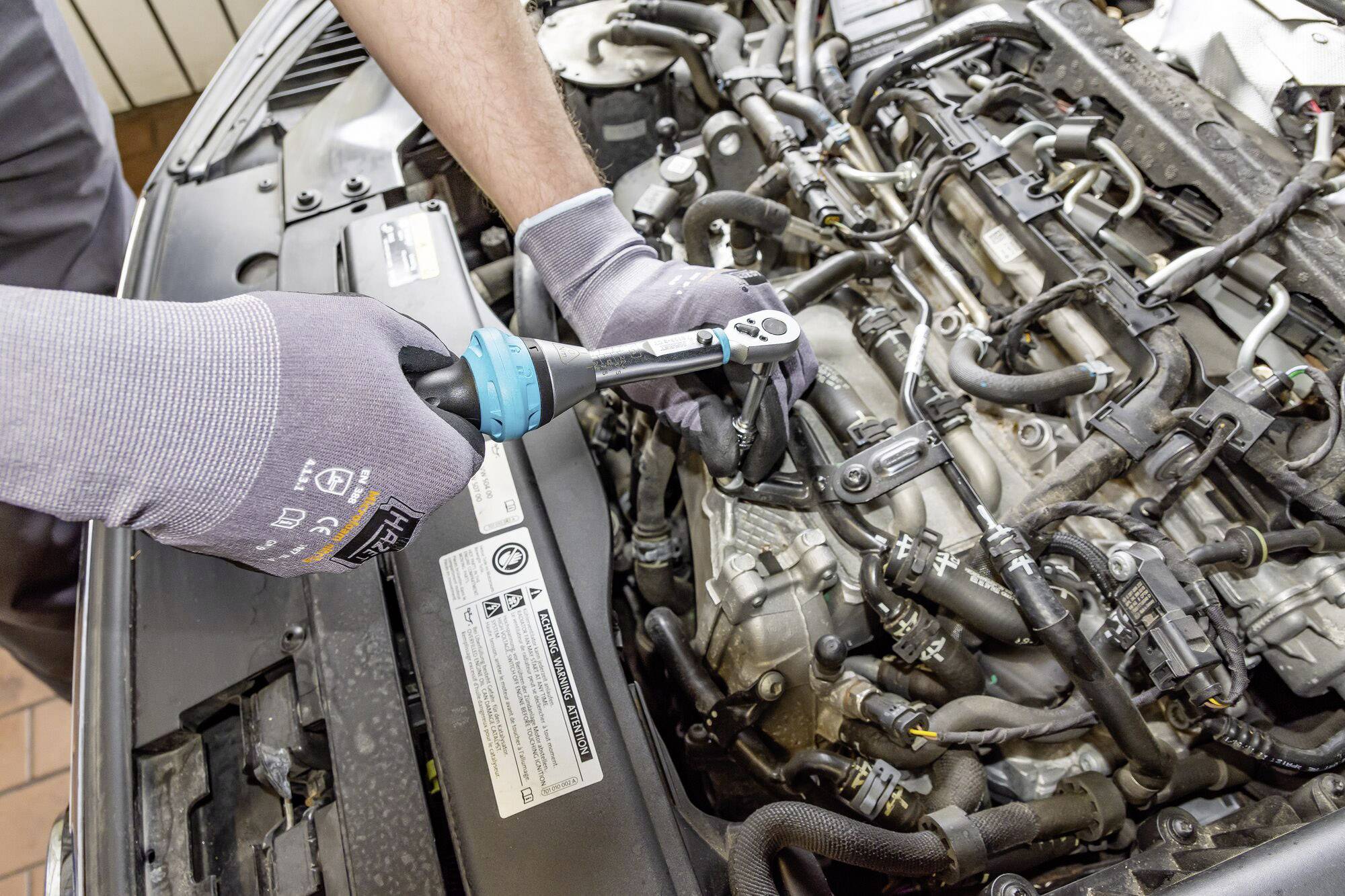 Une personne répare le moteur d'une voiture à l'aide d'une clé dynamométrique positionnée sur une vis du moteur. Le mécanicien porte des gants.