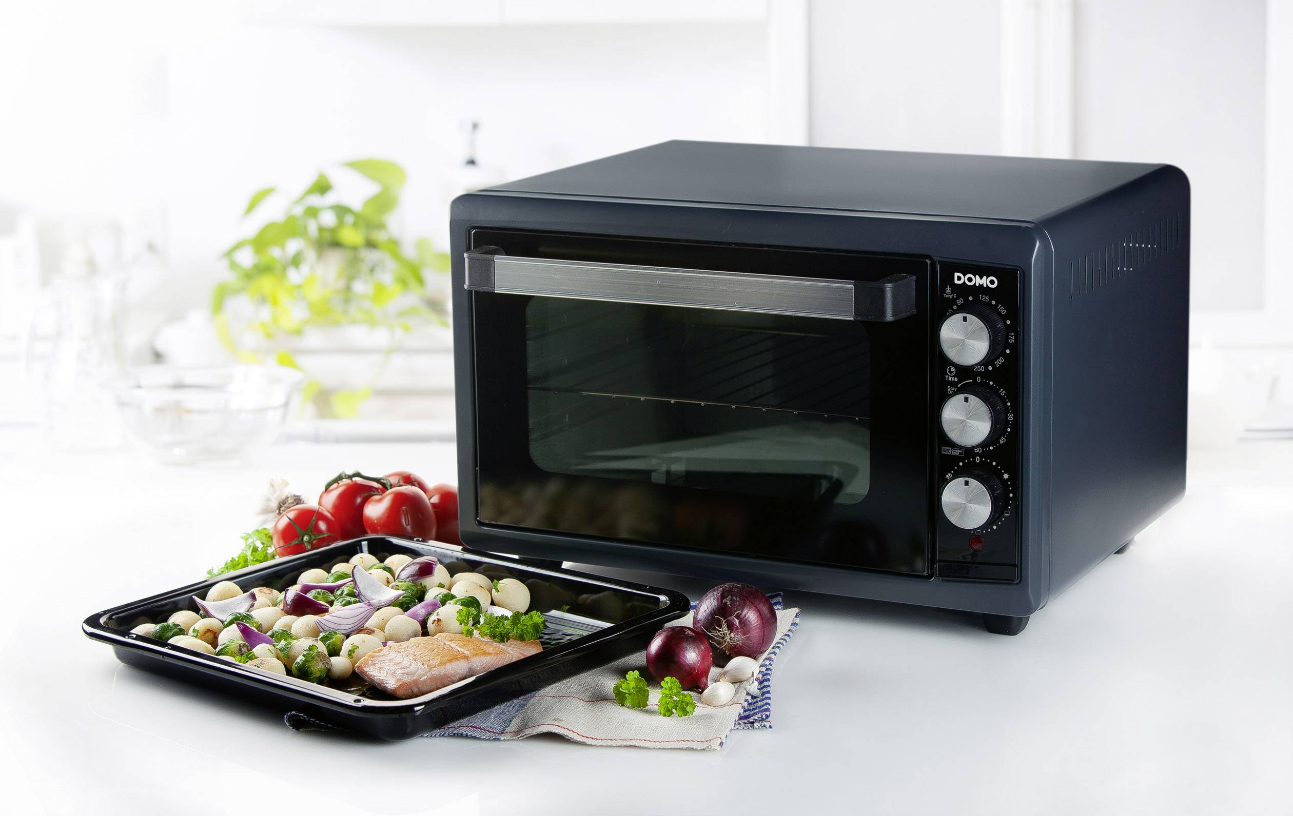 Un four mini noir est posé sur un plan de travail de cuisine. Devant lui se trouve une plaque de cuisson avec des légumes et du saumon.