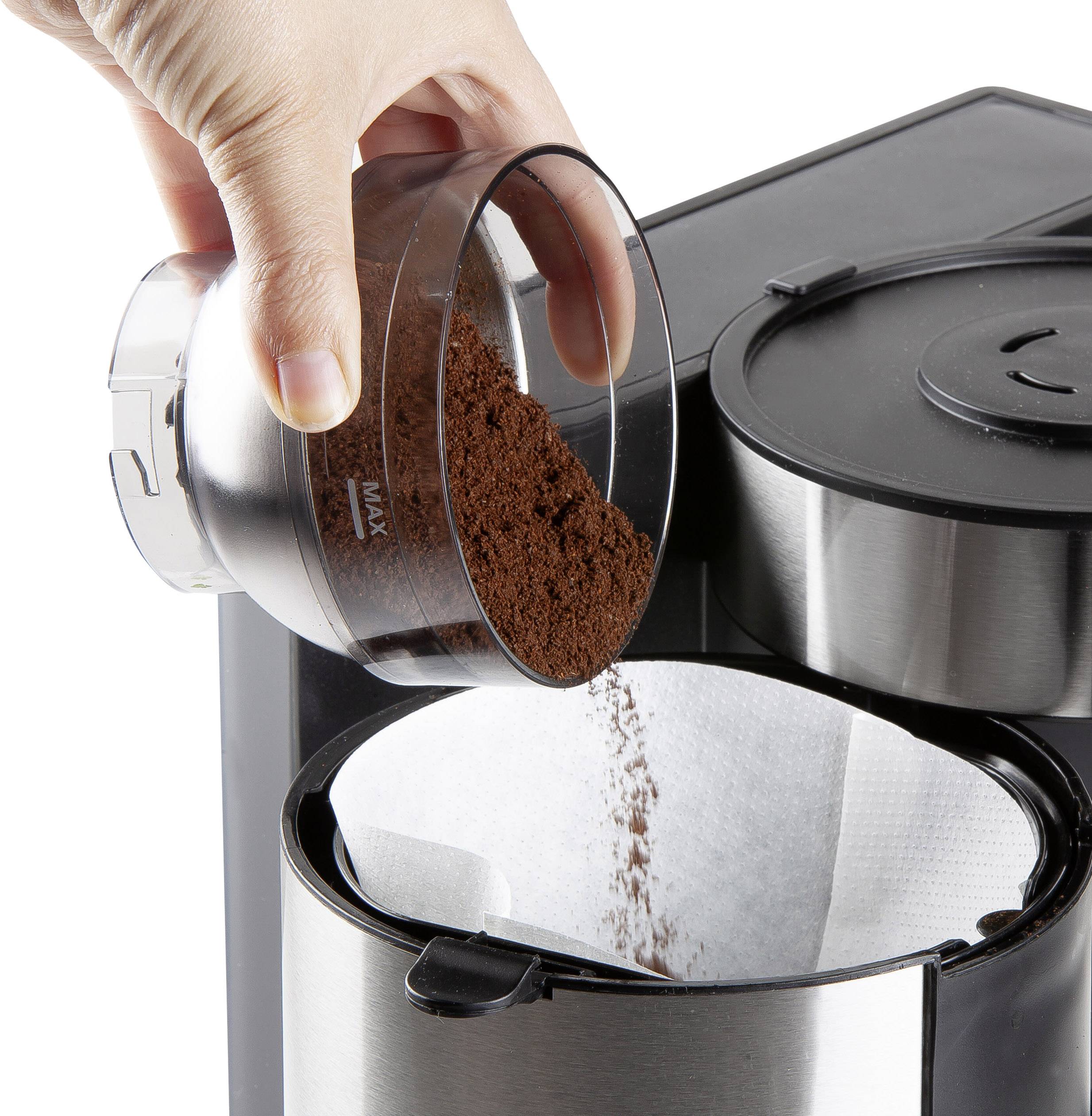 Une main verse du café moulu dans une machine à filtre. Le filtre est ouvert en haut, et la machine est en acier inoxydable et noire.