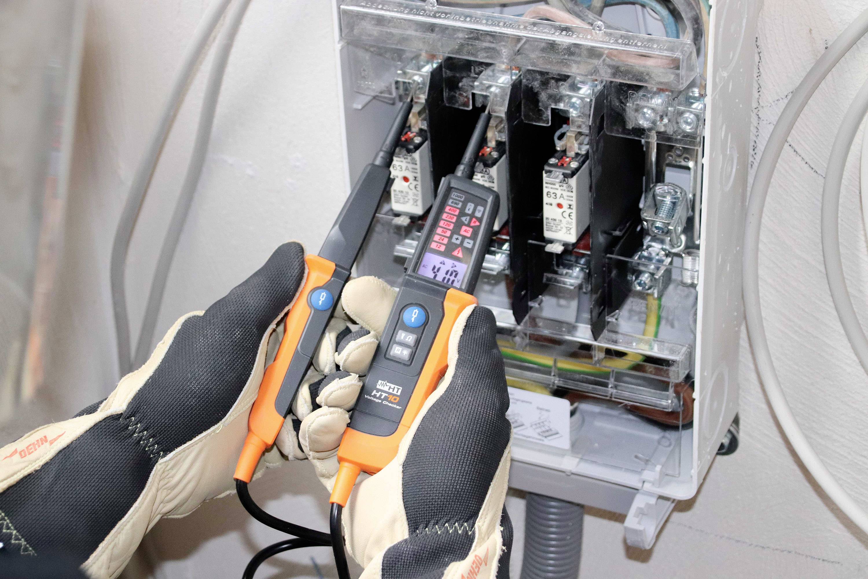 Un électricien porte des gants et utilise un détecteur de tension sur un boîtier de fusibles ouvert.
