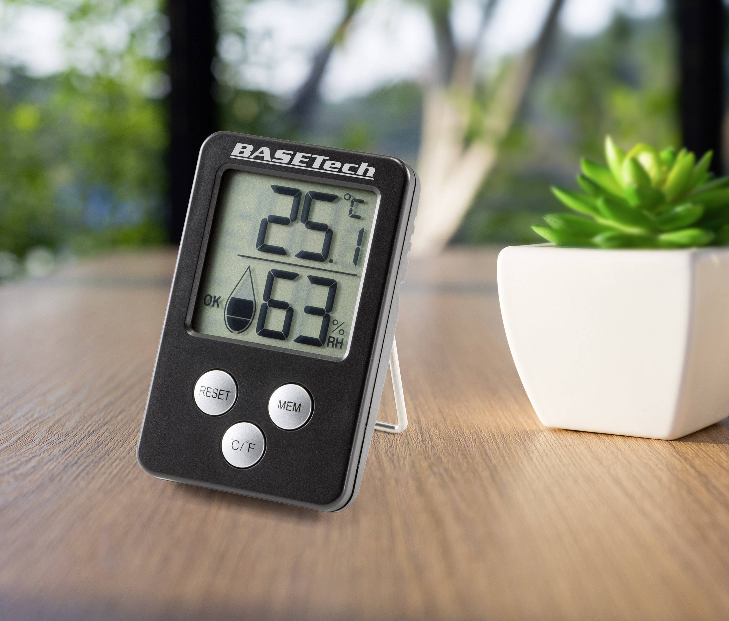 Un thermomètre et hygromètre numérique posé sur une table indique 25,1 degrés Celsius et 63% d'humidité. À côté se trouve une petite plante.