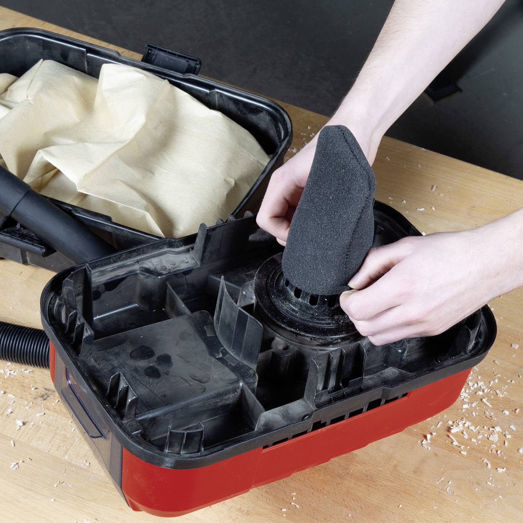 Une personne change le filtre d'un aspirateur. L'aspirateur est ouvert, et le filtre usagé est visible sur une table en bois.