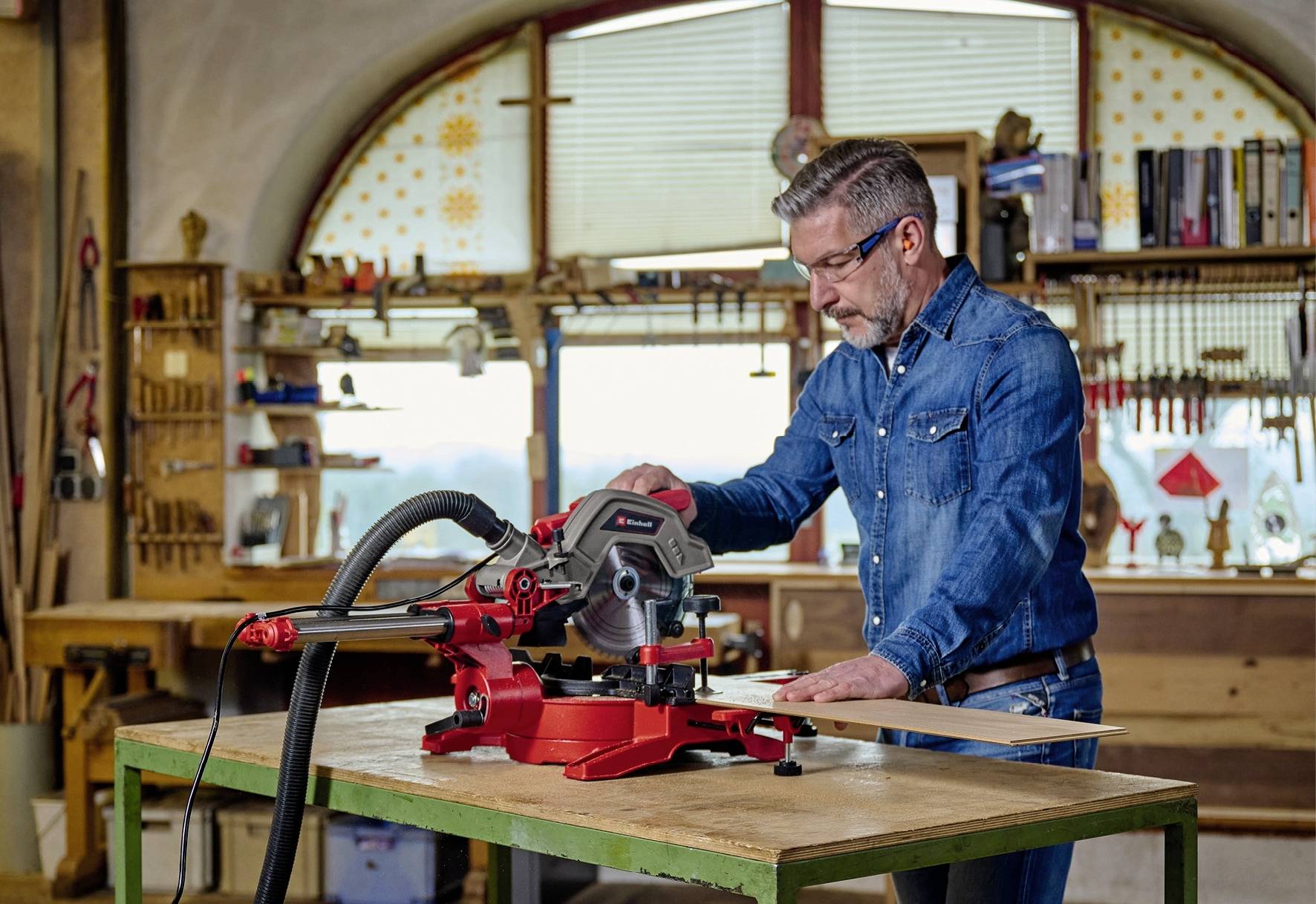 Un homme vêtu de jeans utilise une scie circulaire de table rouge dans un atelier avec un inventaire de bois et des étagères à outils en arrière-plan.